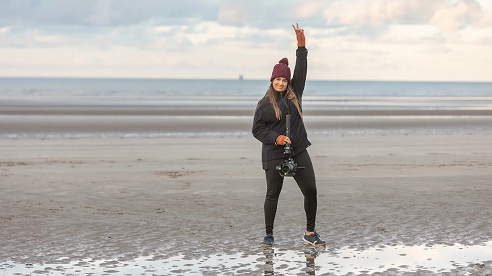A Video Producer standing on a beach holding a camera.