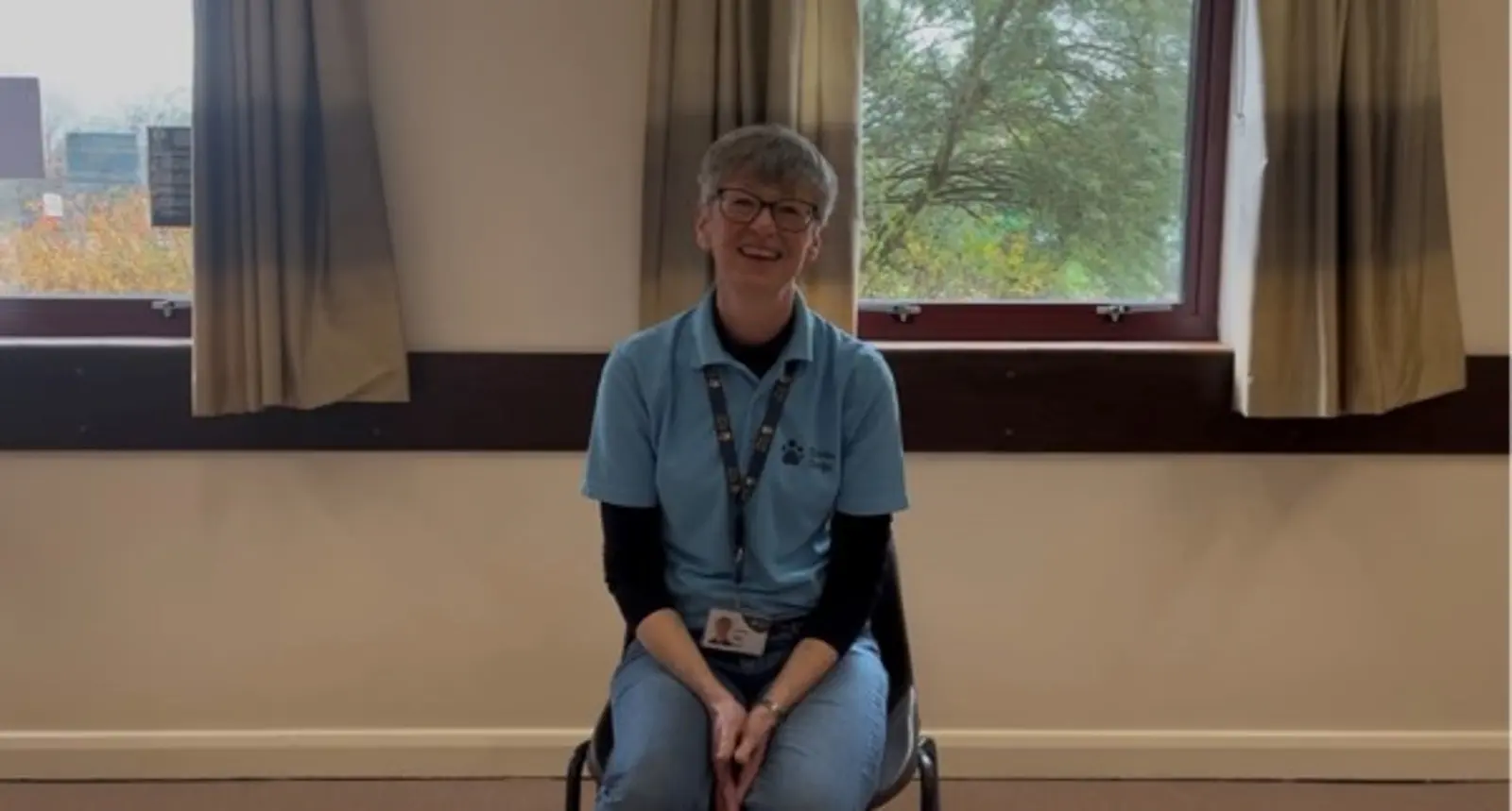 This photo shows Helen, a volunteer puppy class helper. Helen is sitting down on a chair, wearing a blue Guide Dogs polo shirt and lanyard. She is looking towards the camera smiling. Behind her there are two windows with beige curtains with a view of trees outside.