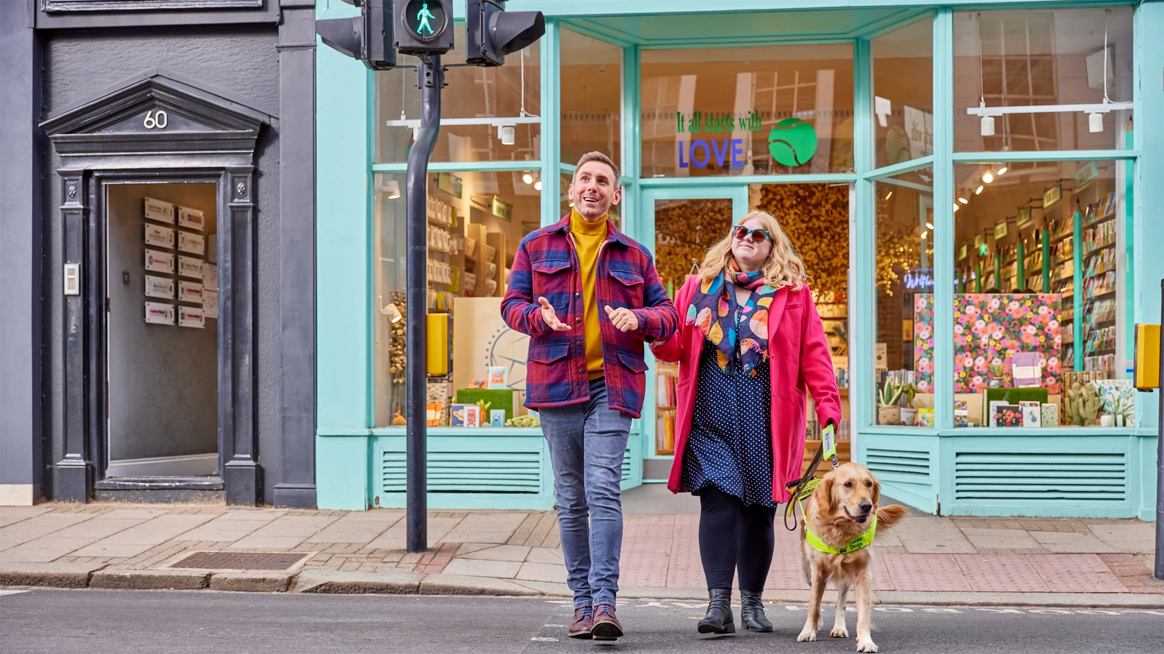 A man guides a woman and her guide dog over a pedestrian crossing, she holds his arm using the C grip.