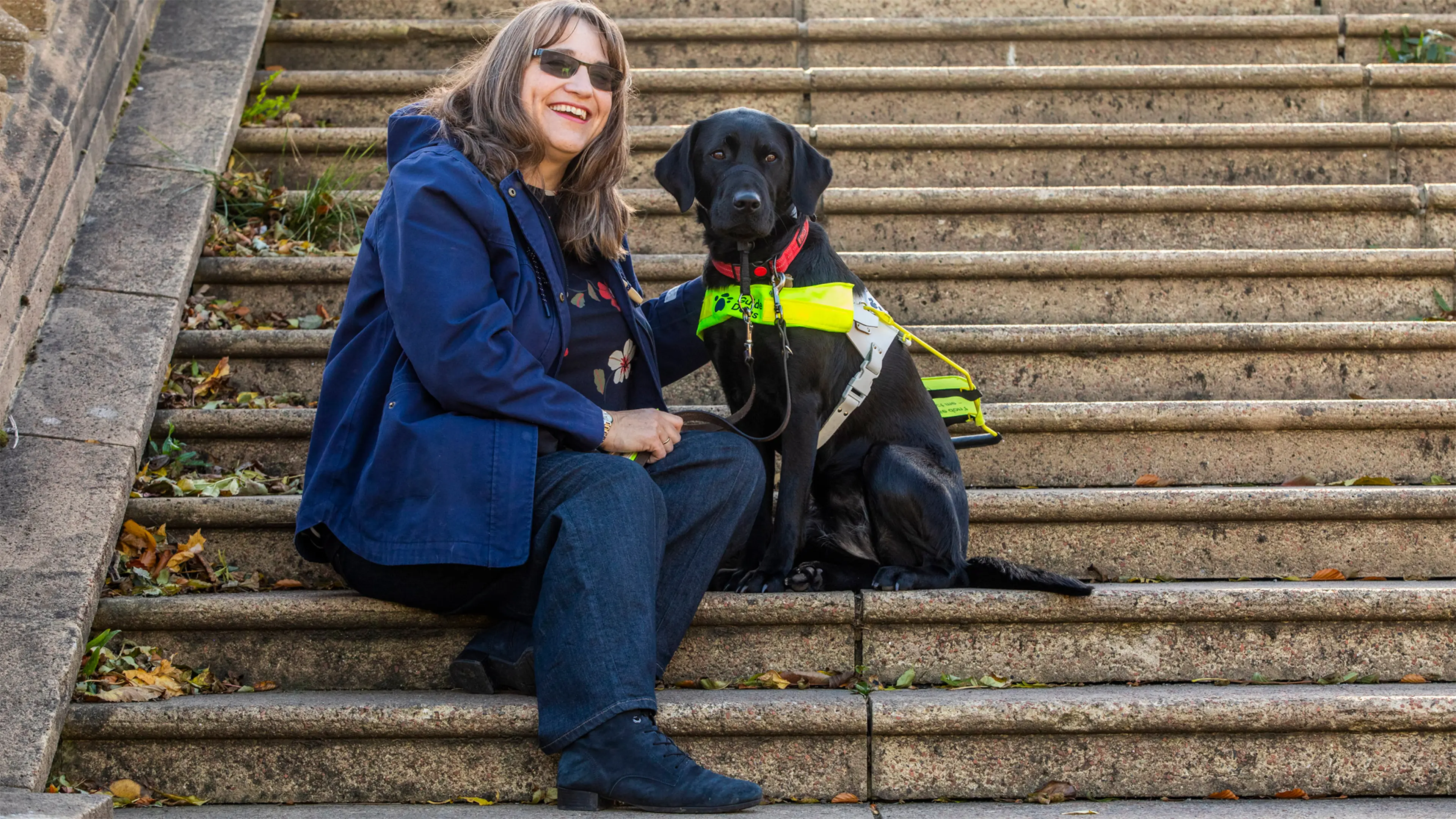 Guide dog owner Lesley sits on some stone steps. Her black Labrador guide dog Kide sits beside her.