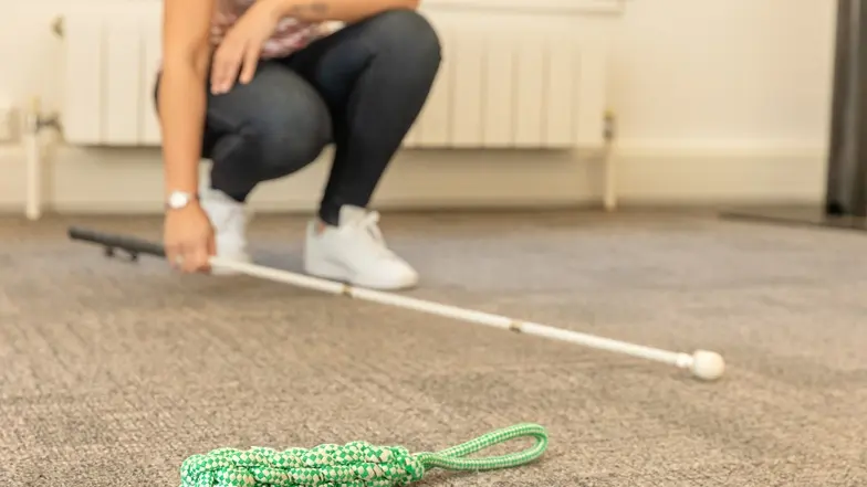 A person crouches down and uses their long cane to  search for a dropped item.