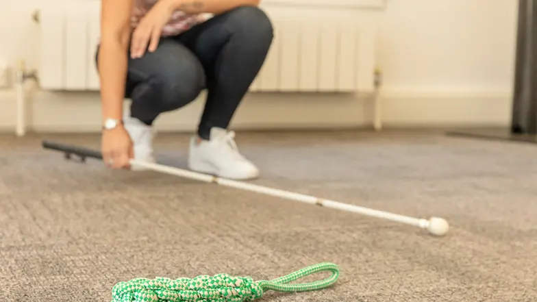 A person crouches down and uses their long cane to  search for a dropped item.