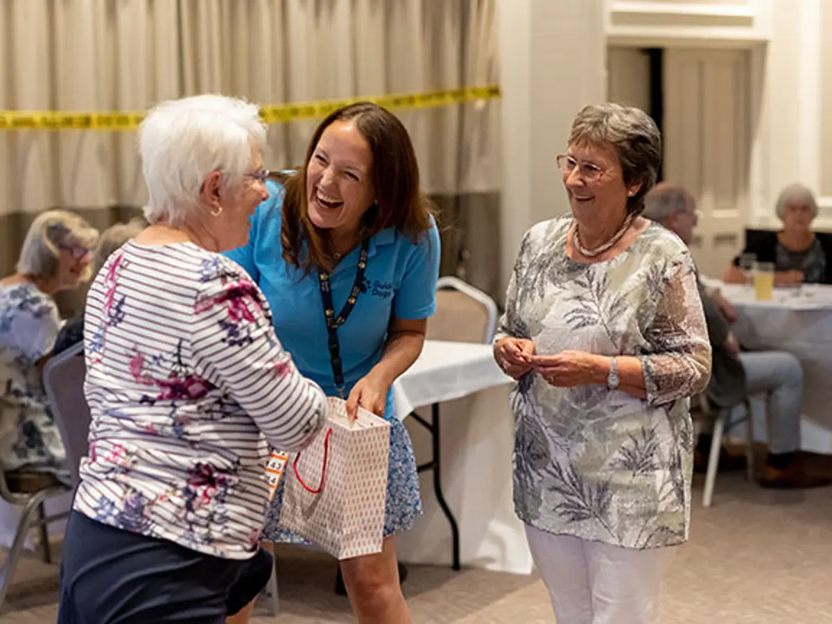 Sue (on the right, wearing a grey top) is at a Guide Dogs event and is laughing with a fellow volunteer and staff member