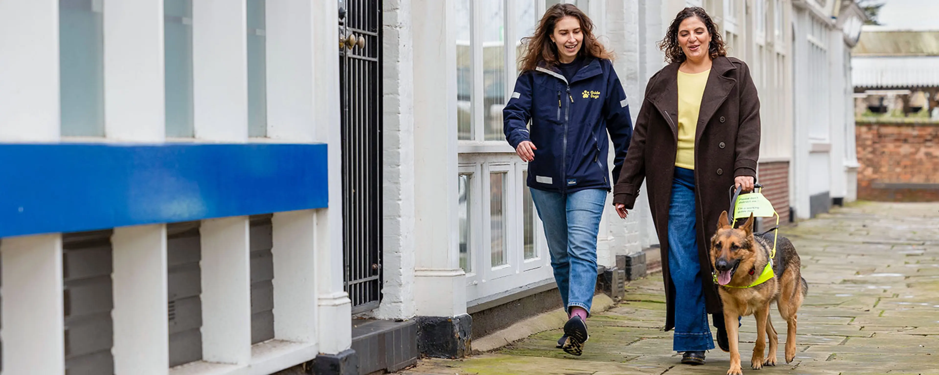 Maria walks with guide dog Pascoe and Guide Dogs Mobility Specialist along a street. 