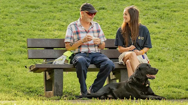 Guide dog owner Alan sits on a park bench with his Guide Dog Mobility Specialist, Angharad and his guide dog, Dane, sits at their feet.