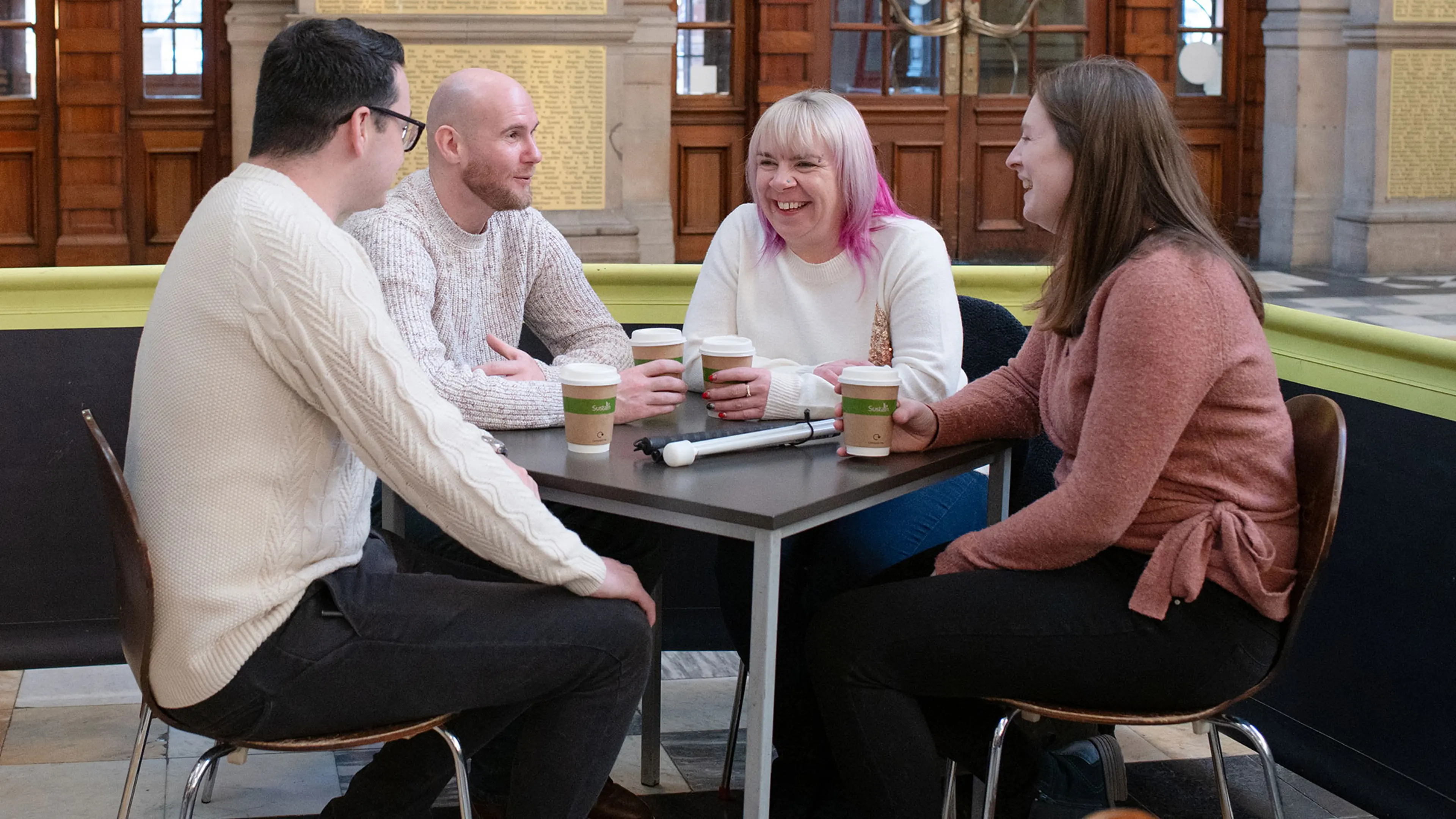 A group of people sitting at a table enjoying a cup of coffee. A white cane is rested on the table.