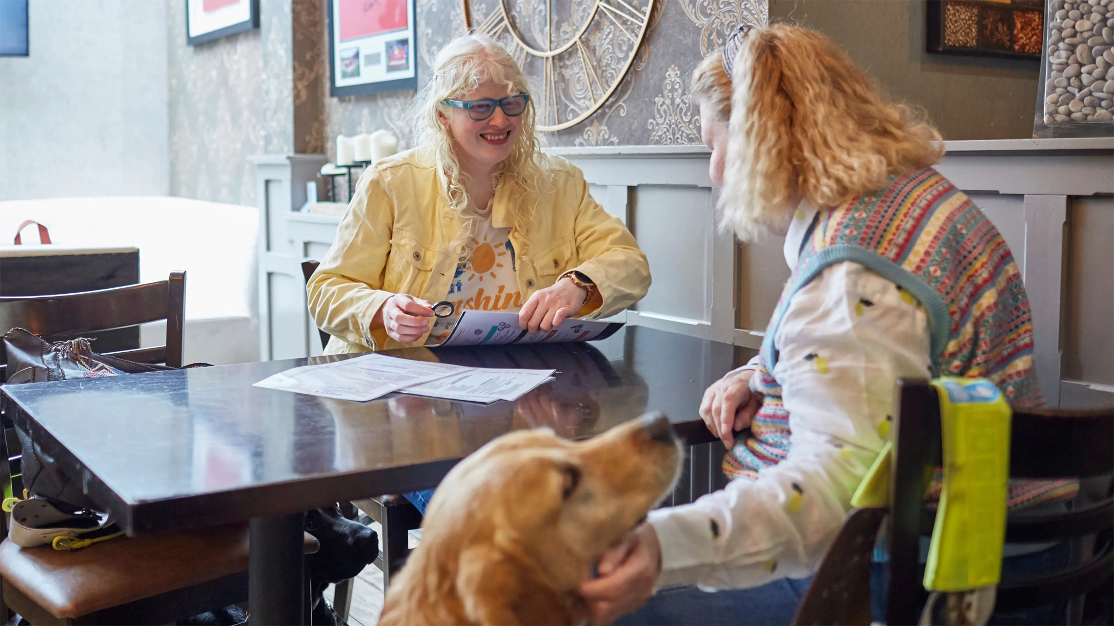 Guide dog owners Karishma and Jane sit at a table in a cafe. Karishma holds a magnifiying glass to read the menu and Jane strokes her guide dog.