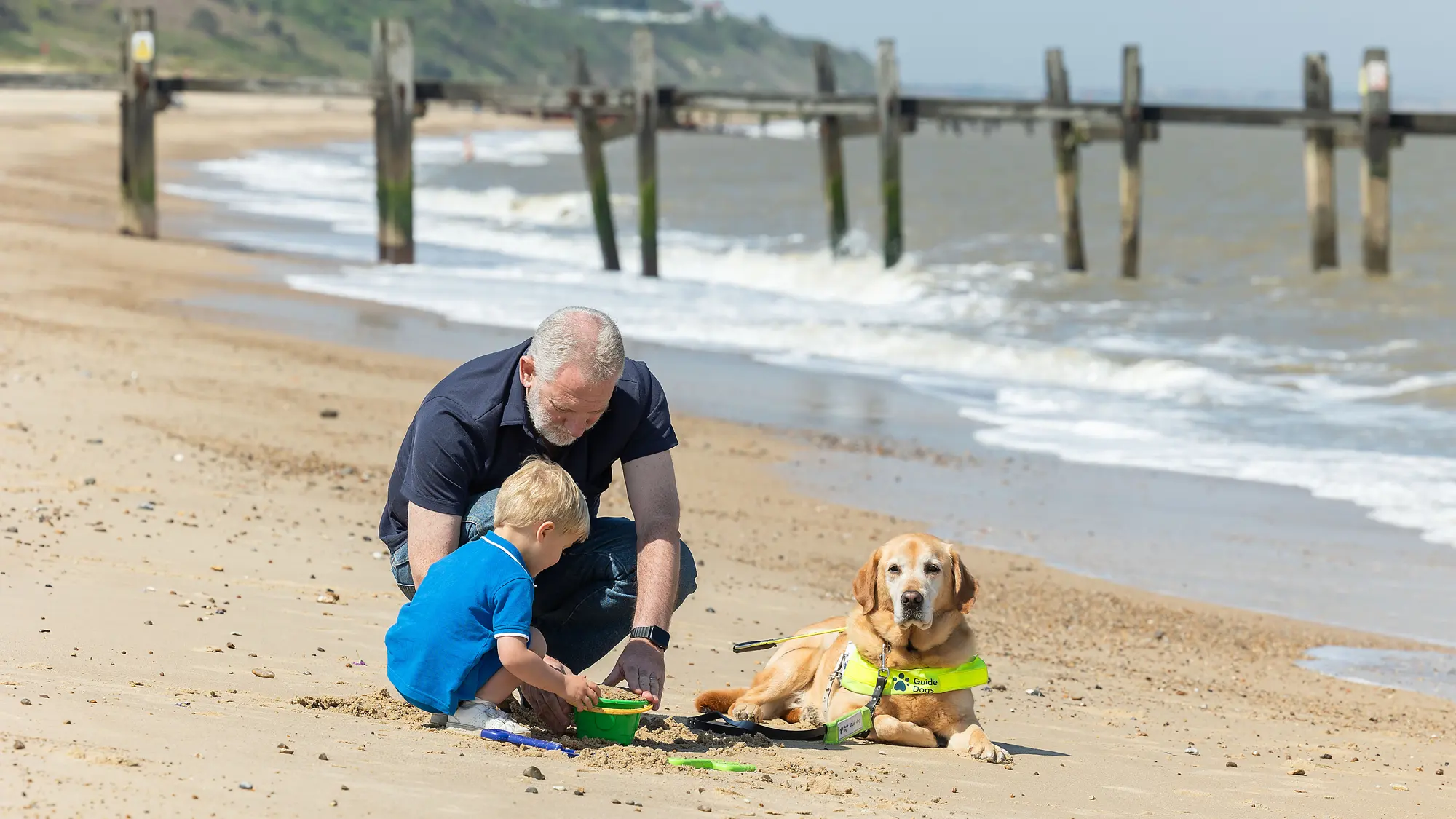 Guide dog owner Simon plays on the beach with his grandson, his guide dog Mayne lies patiently beside them.