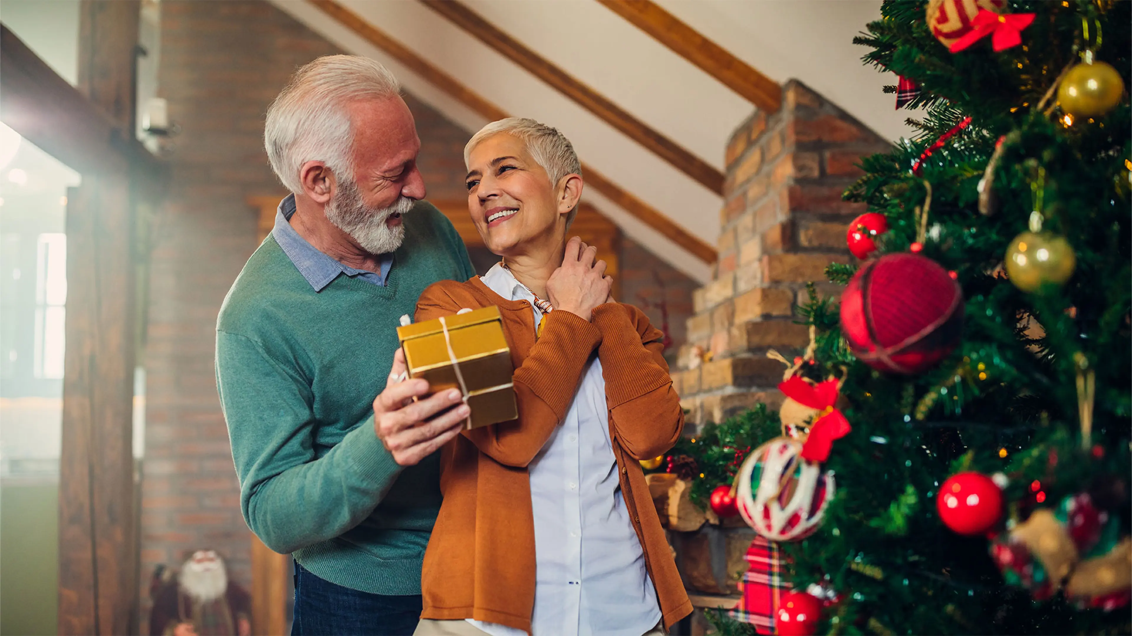 A man holds a gold box in his hand and looks towards a smiling woman in front of a Christmas tree.