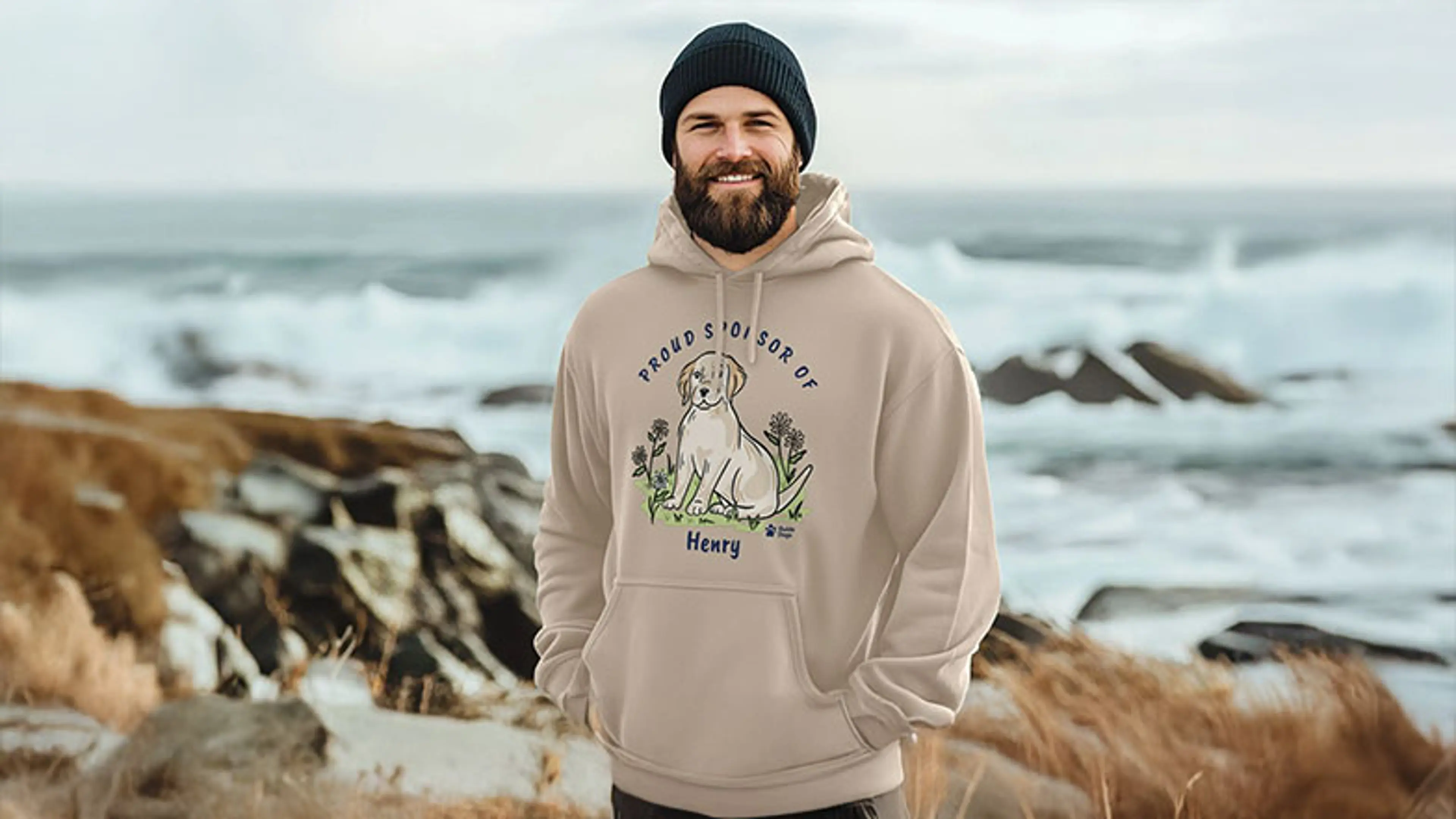 A bearded man wearing a 'Proud sponsor of Henry' sweatshirt standing on a beach the sea in the background.