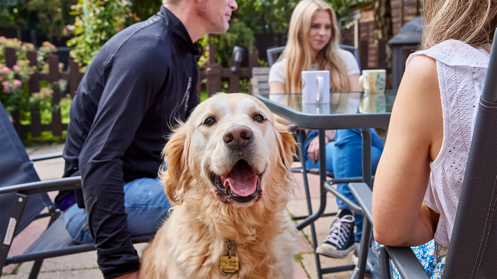 Guide dog owner Scott and his family sit at a garden table, his guide dog Milo looks straight at the camera with his tongue out.