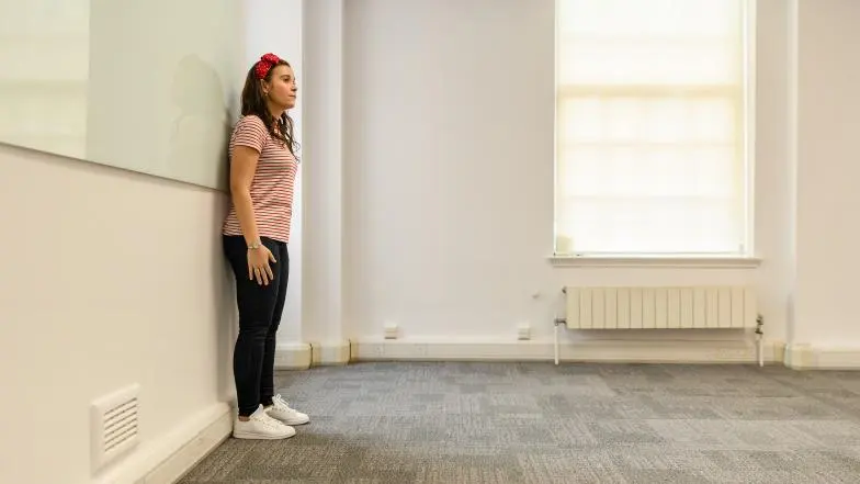 A woman stands against a wall in a meeting room, demonstrating the squaring off technique.