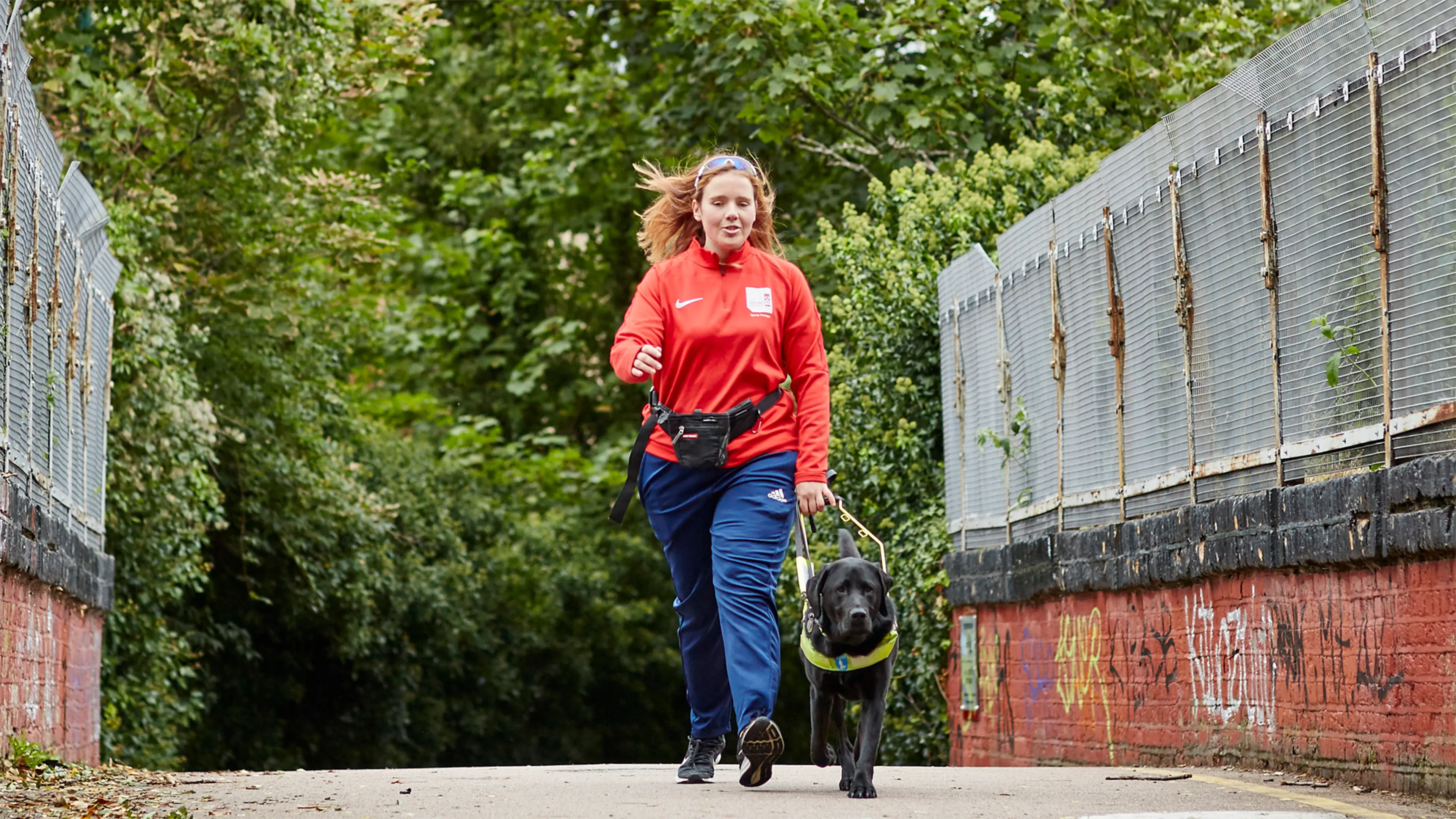 Lois, a guide dog owner, strides over a bridge holding the harness of her guide dog, black Labrador Buster.
