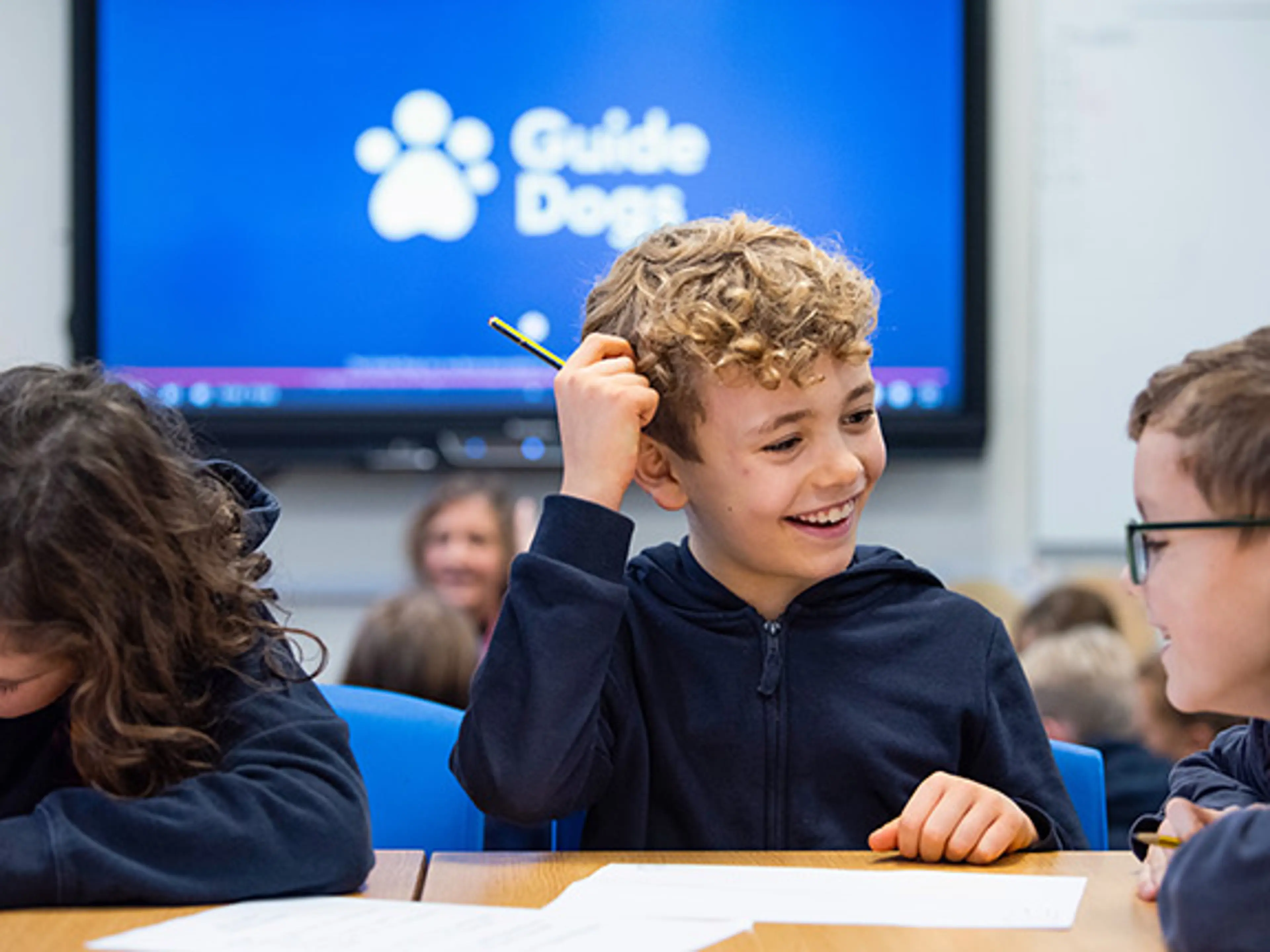 School children in a classroom talking and completing a worksheet while sat in front of a large screen that has the Guide Dogs logo.