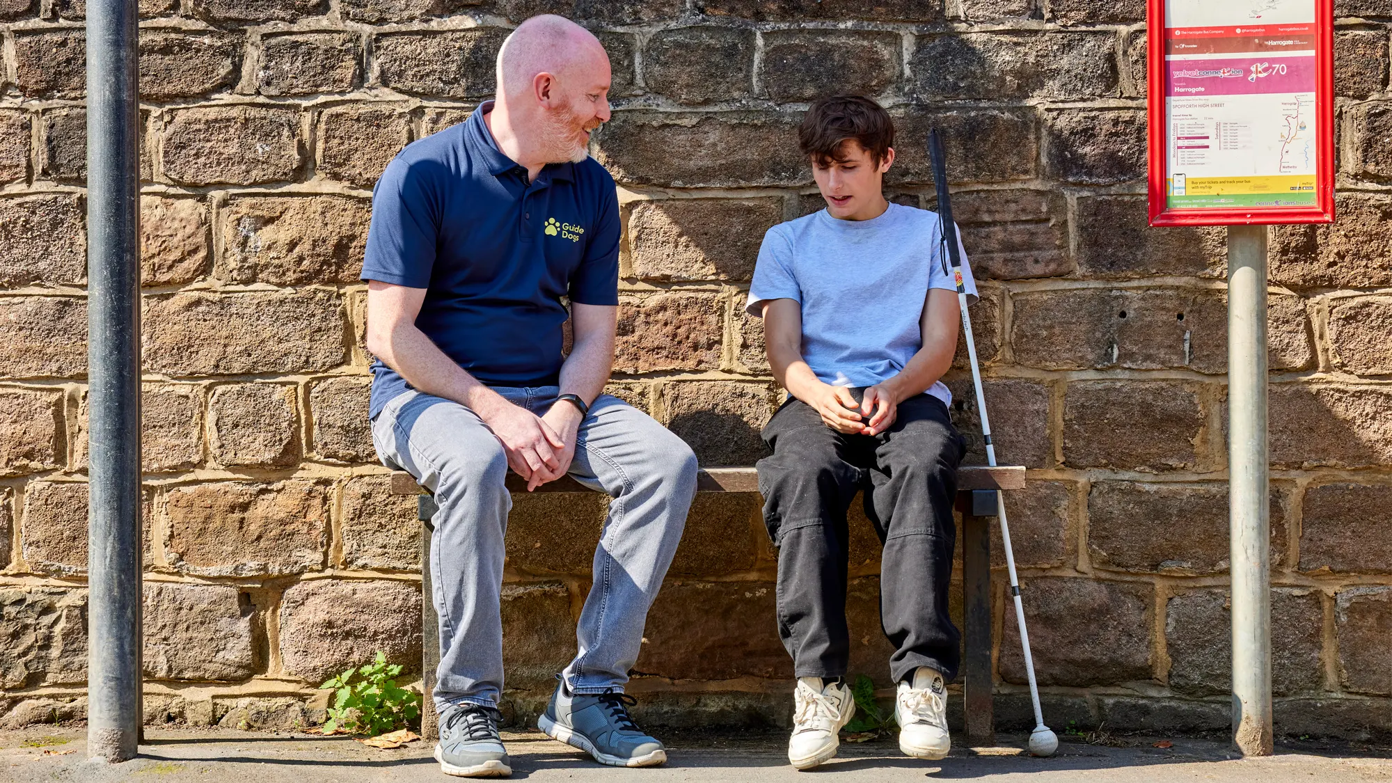 A teenager with sight loss sits talking with his Guide Dogs Habilitation Specialist on a bus stop bench.