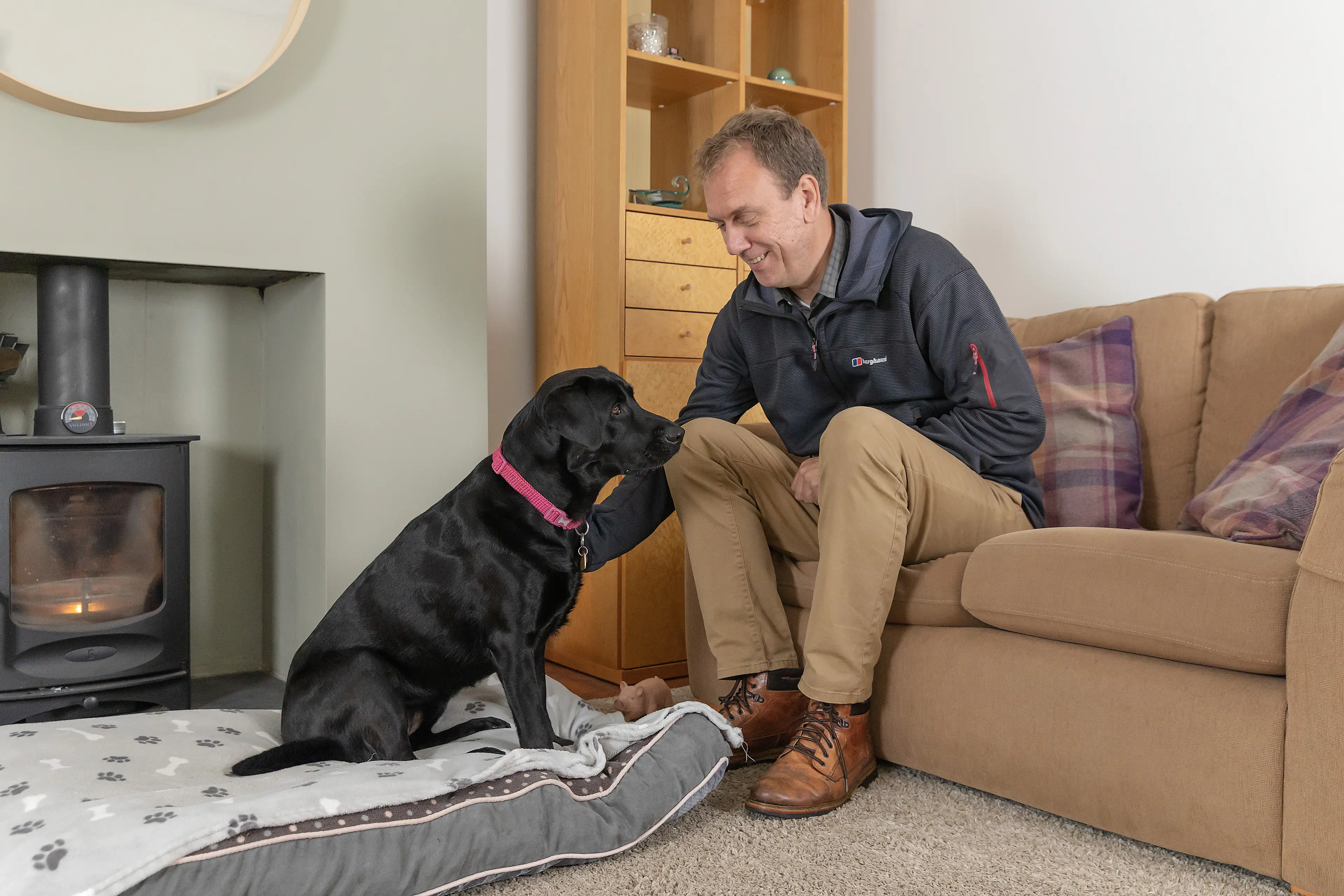A volunteer fosterer sits on his sofa beside a black Labrador guide dog in training
