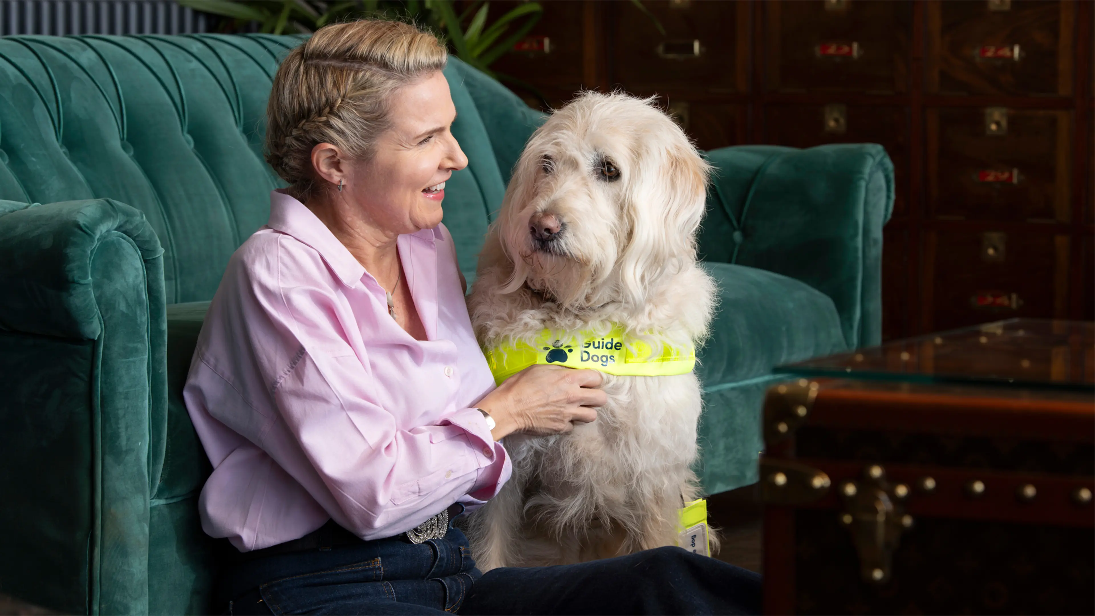 Guide dog owner Verity sits on the floor with her back against a green sofa next to her guide dog, Labradoodle Luna.