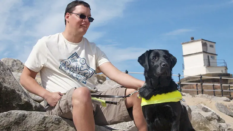 Man with black Guide Dog sat on rocks on beach.