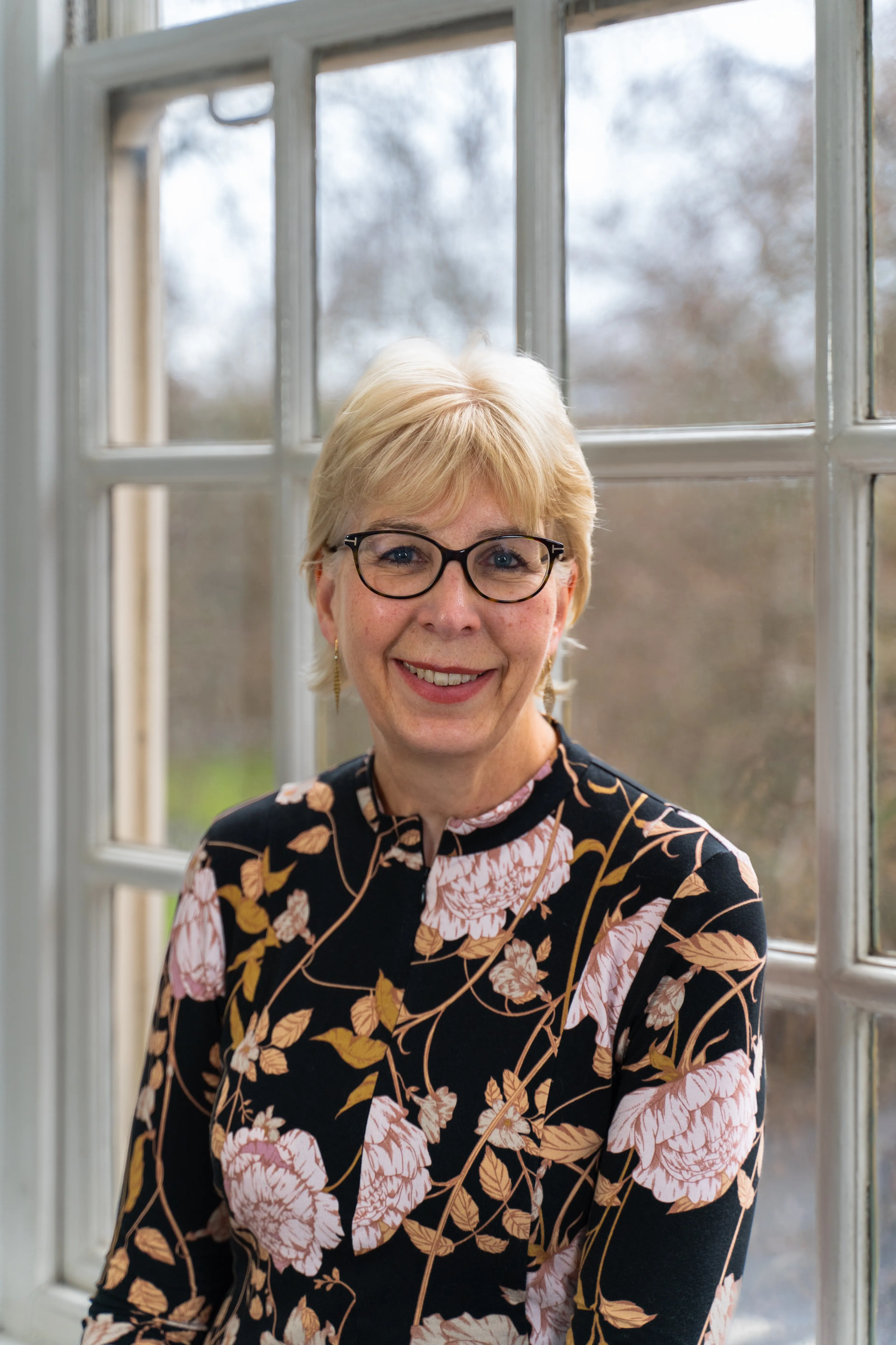 Isabel Hudson smiles at the camera. She is sitting by a window, wearing glasses and a floral top.