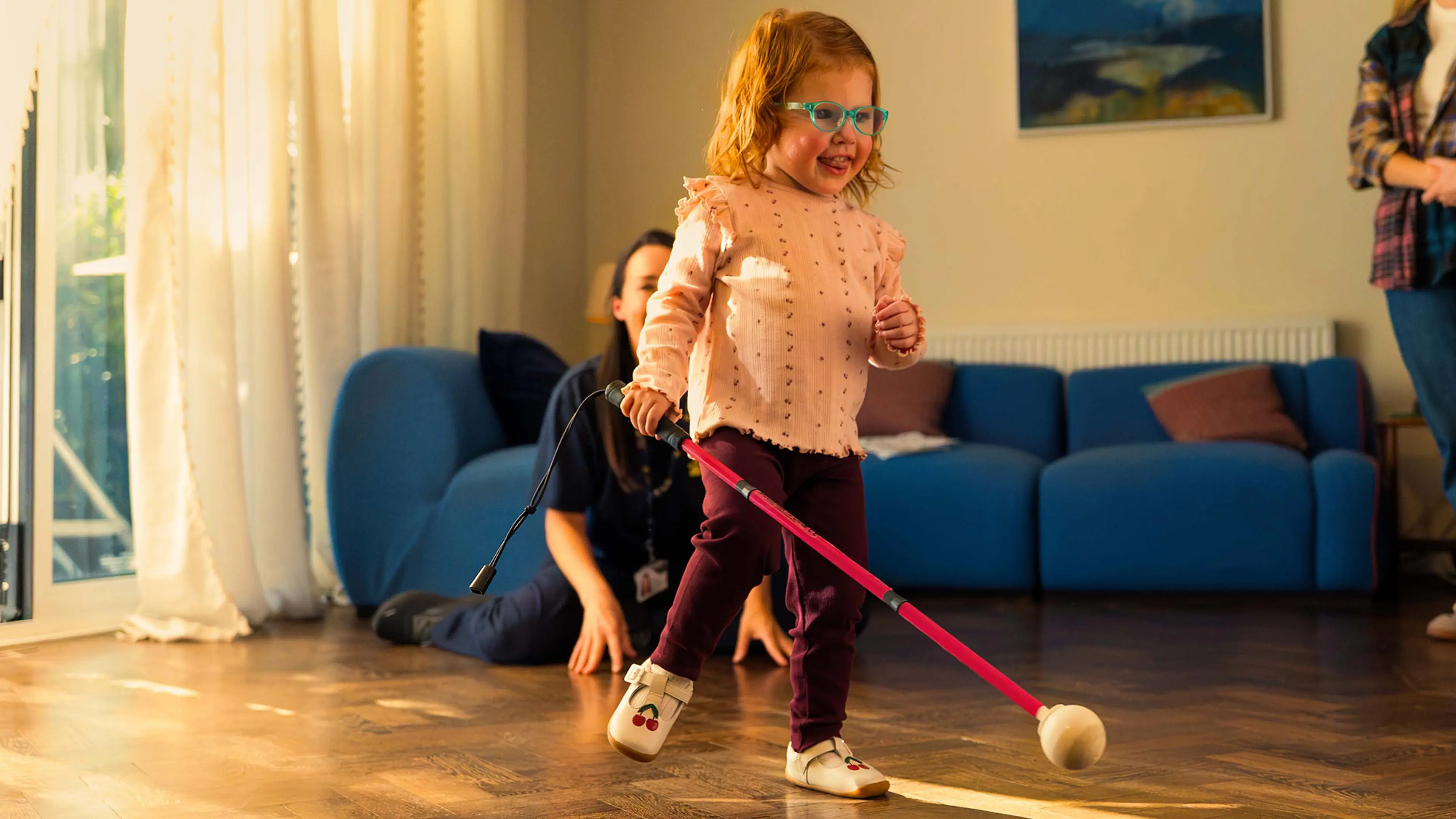 Martha, a child with a vision impairment, is walking through her home using her long cane. She is supported by her Habilitation Specialist, Lisa.