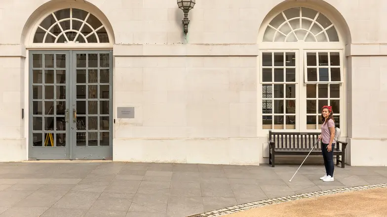 A woman stands off to the right with a long cane, ready to walk across a courtyard.