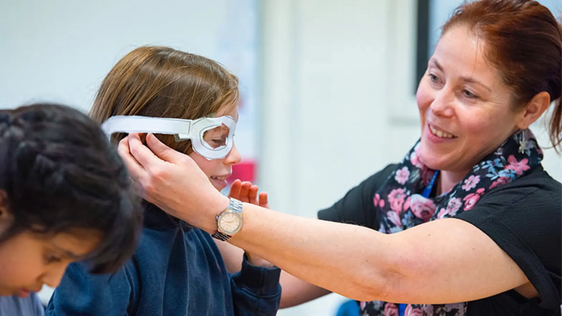 A teacher helps a child try on paper cut-out glasses frames.