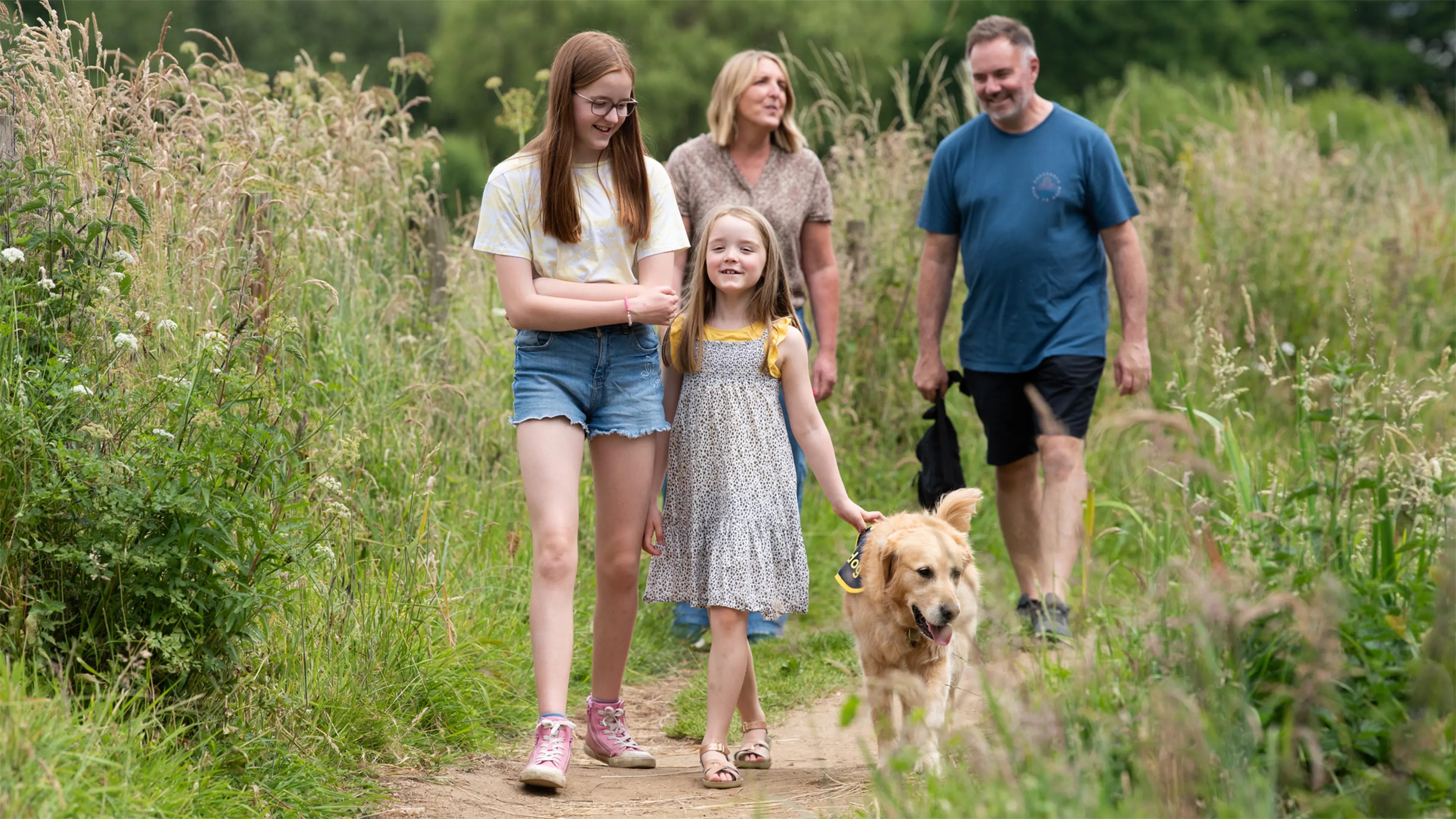 Freya, a young person with vision impairment, walks through a field of tall grass with her buddy dog Gwen and her family.
