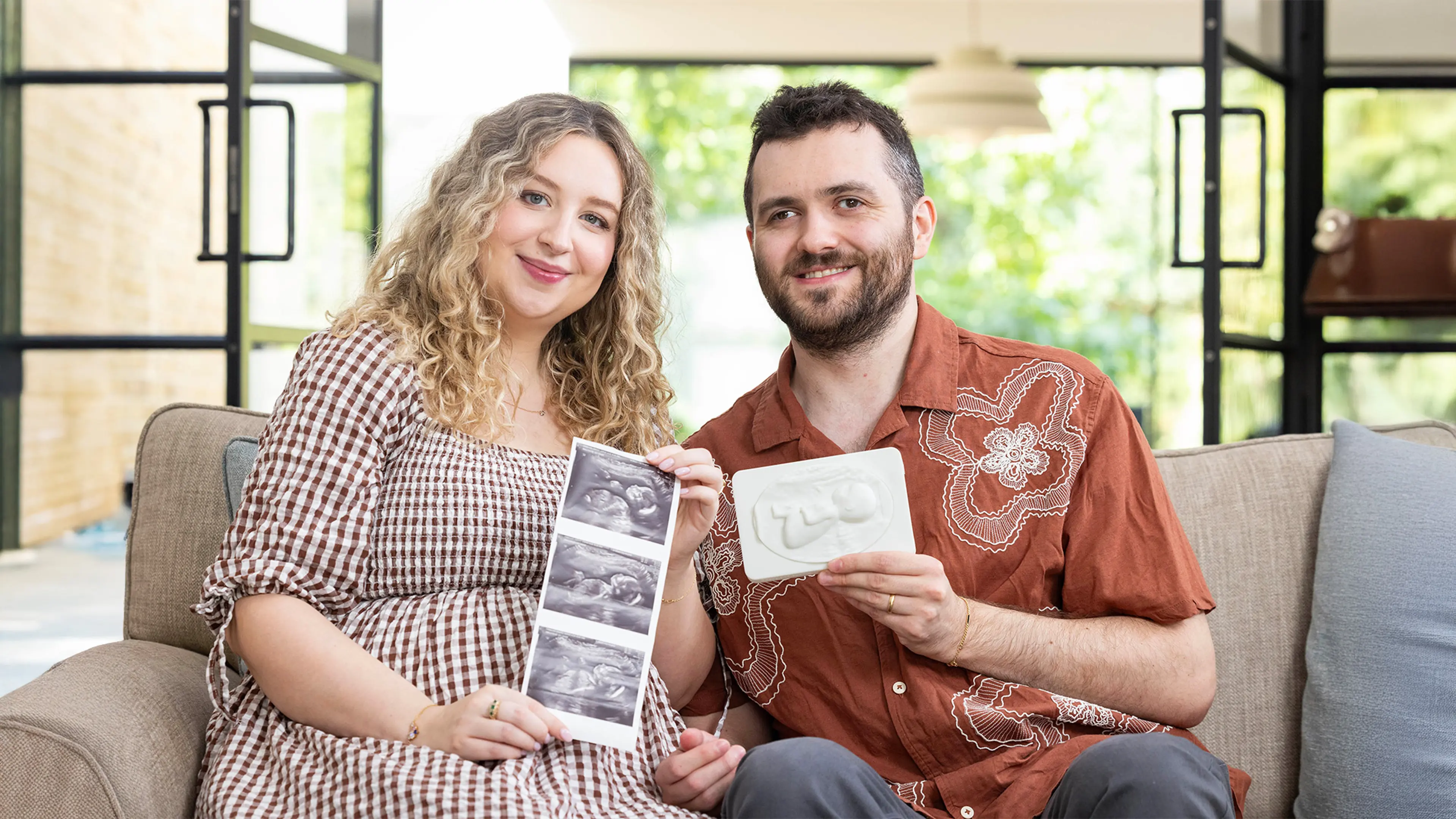 Sophy and Reece sit on the sofa smiling as they look at their First Hello tactile sonogram of their baby. Sophy is holding their 2D ultrasounds scans towards the camera while Reece traces the details of their tactile sonogram for the first time.