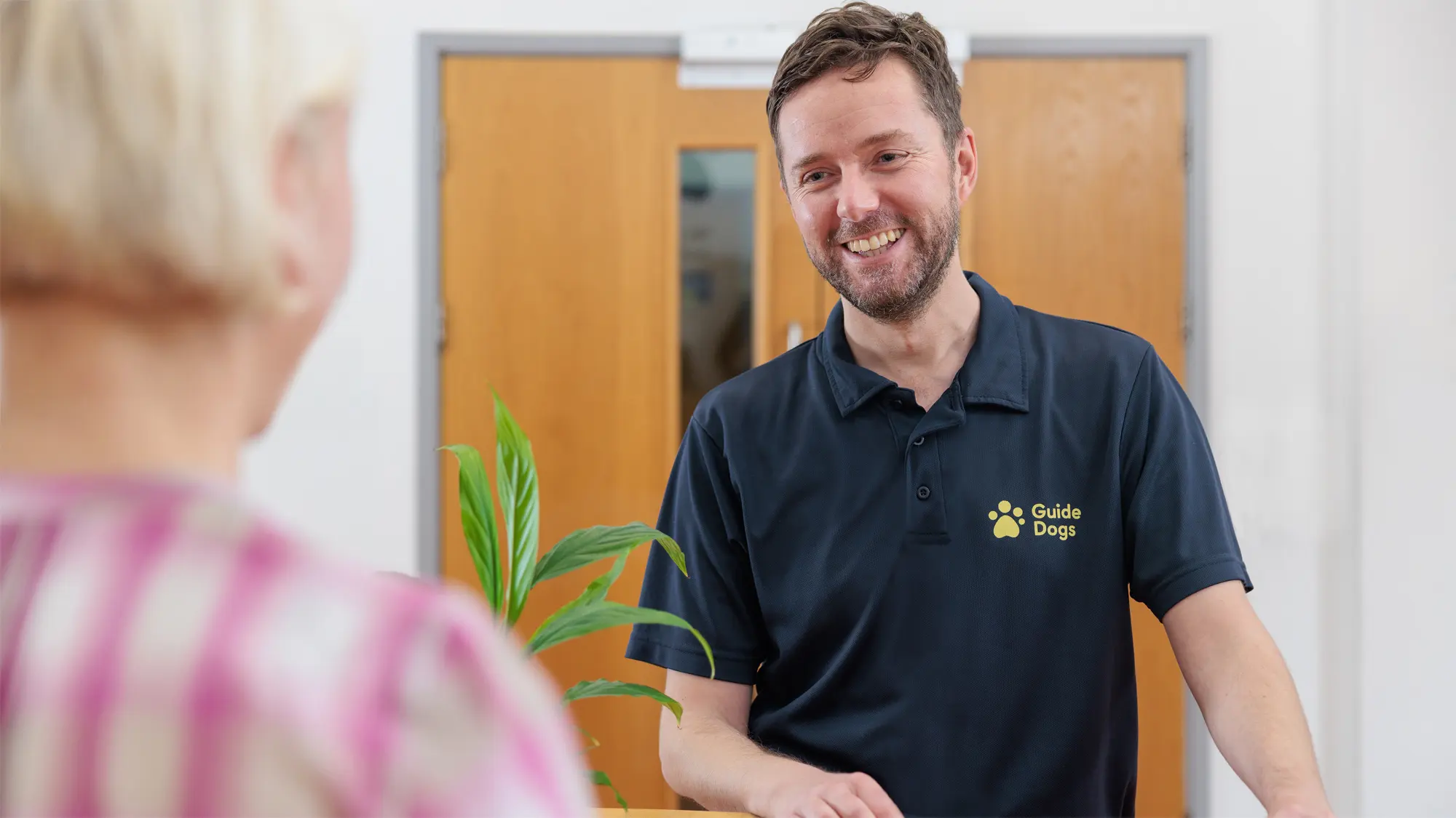 A Guide Dogs employee stands at a reception desk in a Guide Dogs branded polo shirt. 