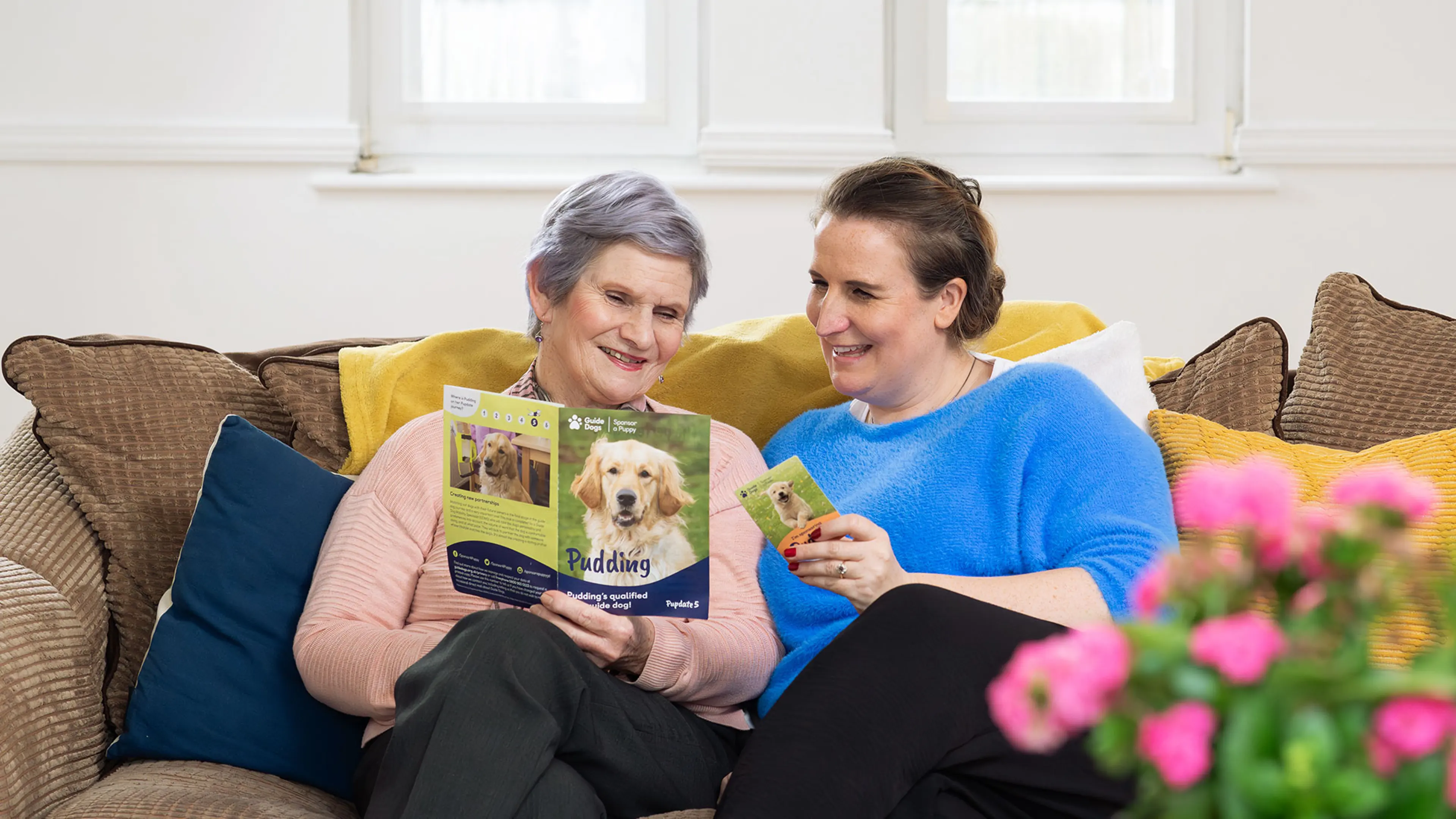 Michelle and her mum sit together on a sofa. Michelle is holding a Pudding fridge magnet and her mum is holding a Pudding printed Pupdate.