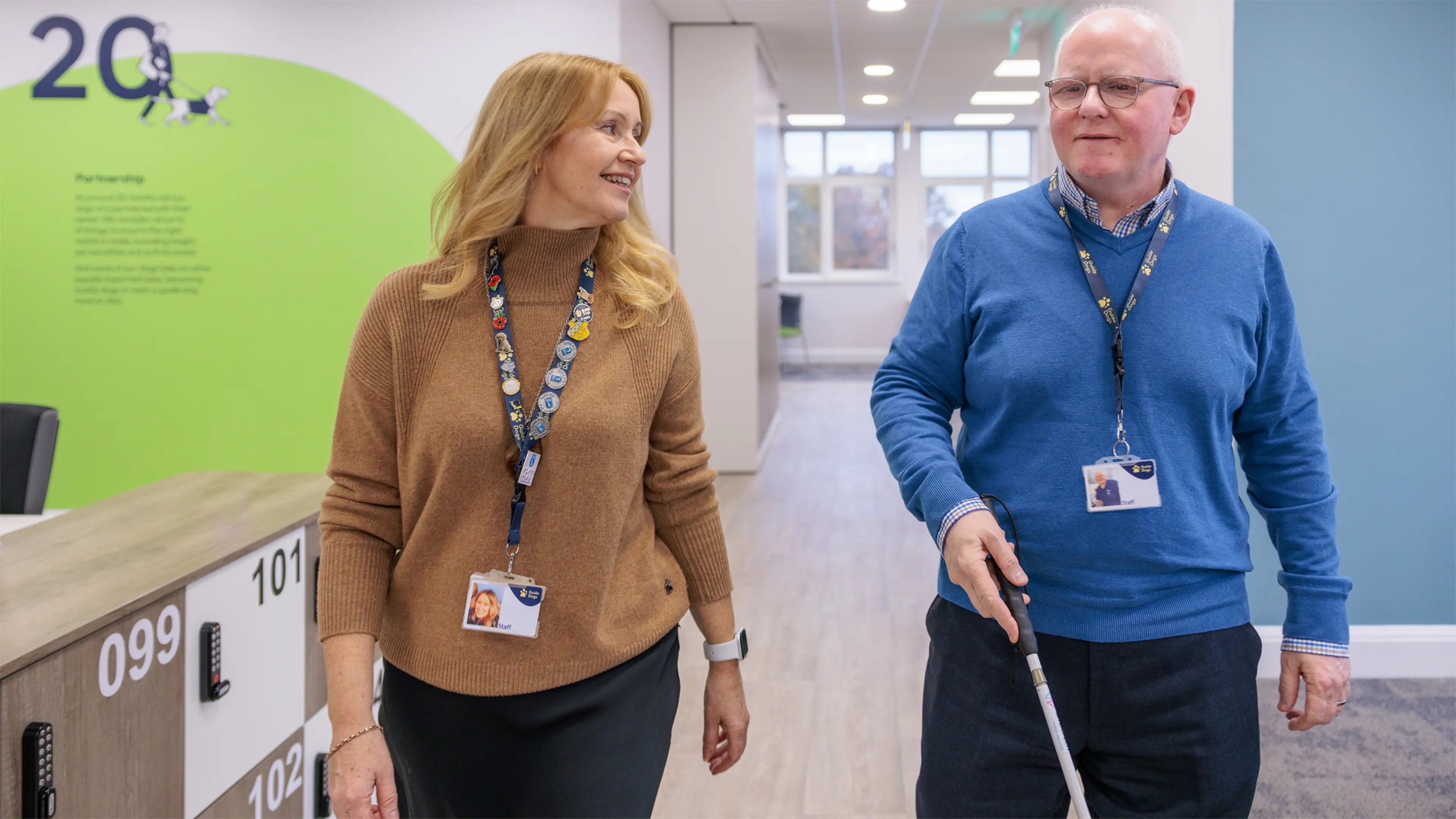 Two Guide Dogs colleagues walk through an office side-by-side, one of them uses a long cane to navigate. 
