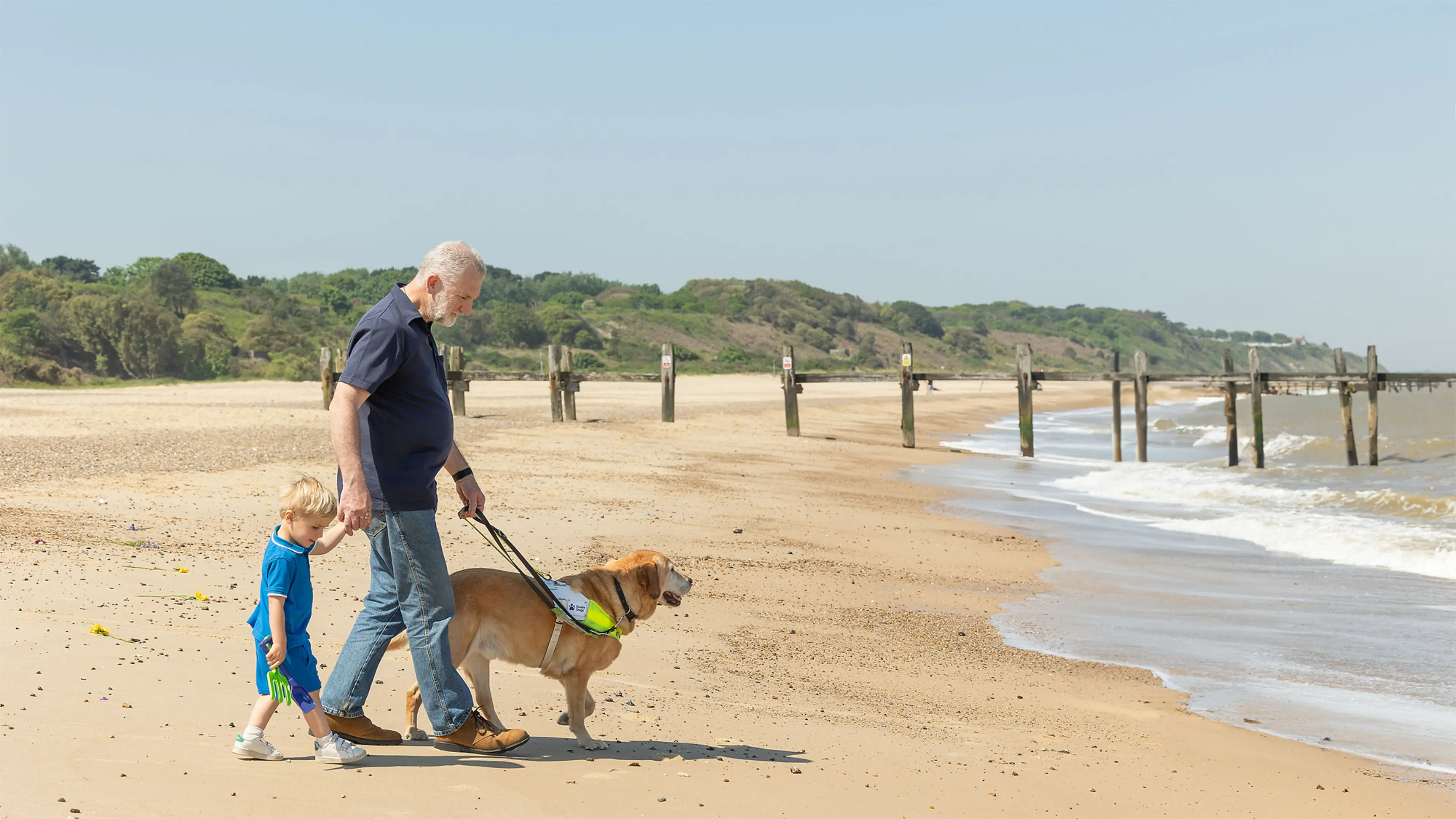 Guide dog owner Simon holds his grandson's hand and his guide dog's harness as they walk on a sandy beach.