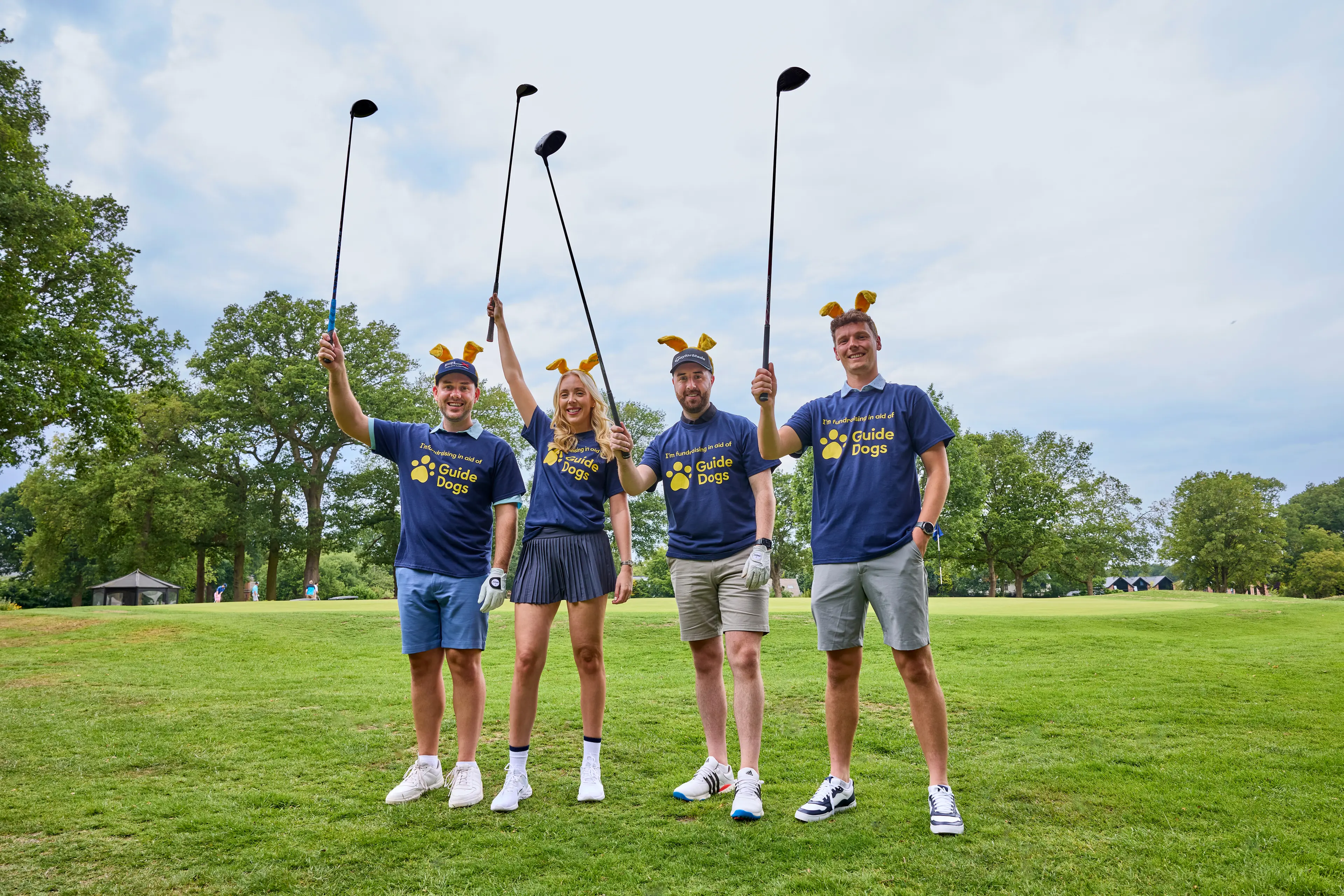 A woman and 3 men stand side by side on a golf course wearing Guide Dogs t-shirts and ears, holding golf clubs in the air.