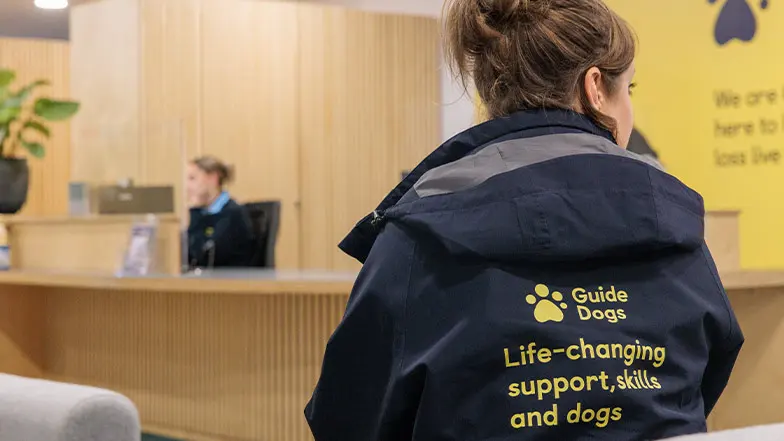 A Guide Dogs staff member approaches a reception desk, her jacket has the Guide Dogs logo and slogan on the back.
