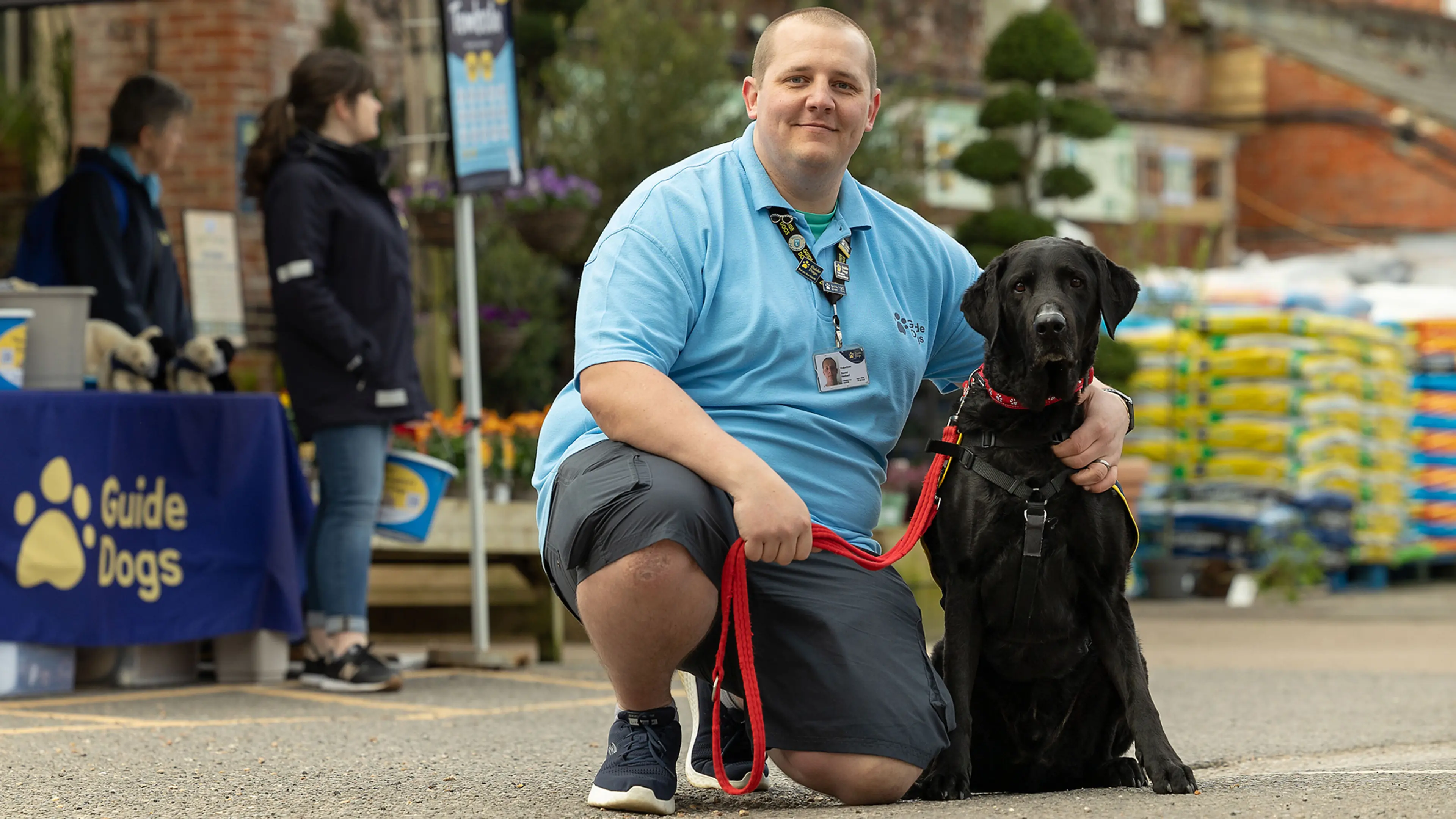 A Guide Dogs Fundraising Group Co-Ordinator kneels in front of a fundraising stall with an ambassador dog.