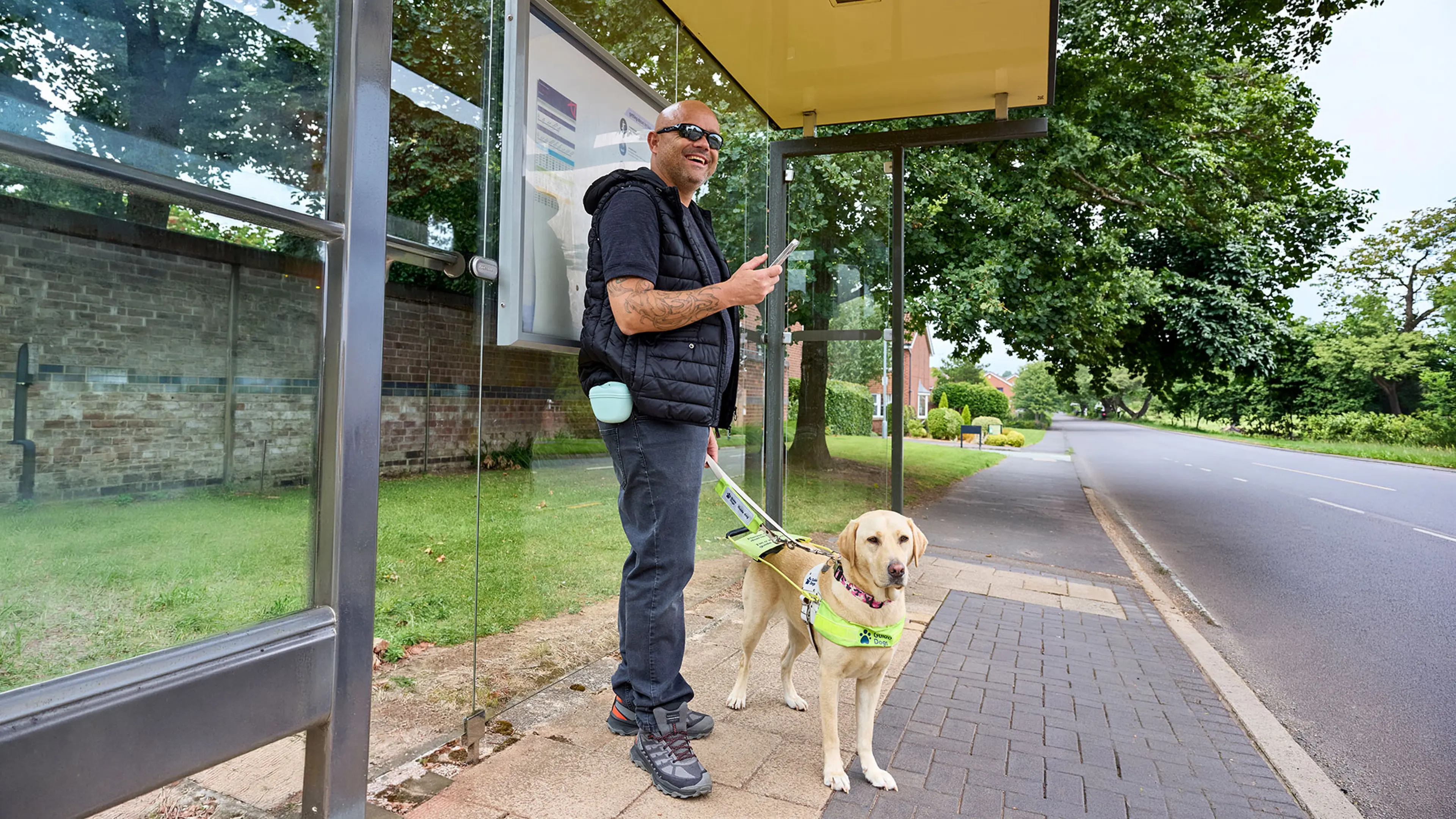 Guide dog owner Wayne smiling and standing at a bus stop with guide dog Liberty. 
