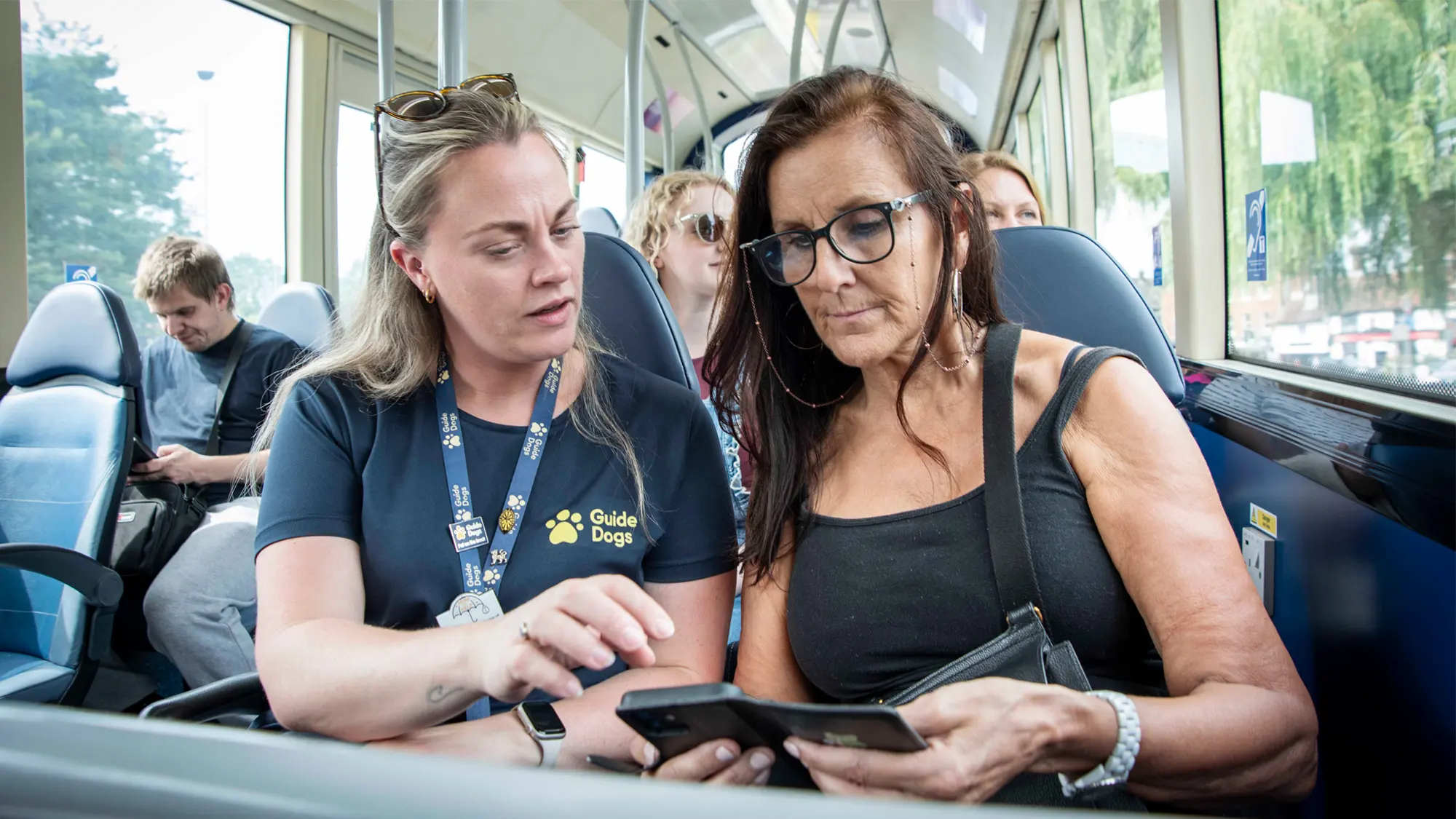 A woman with vision impairment sits on the bus with a Vision Rehabilitation Specialist, as they look at her phone together.