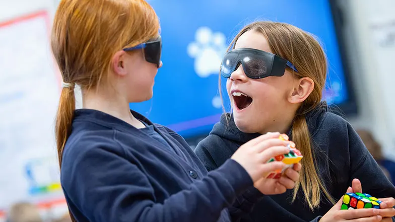 Two young girls in a classroom wearing simulation spectacles and doing a tactile Rubik’s cube activity.