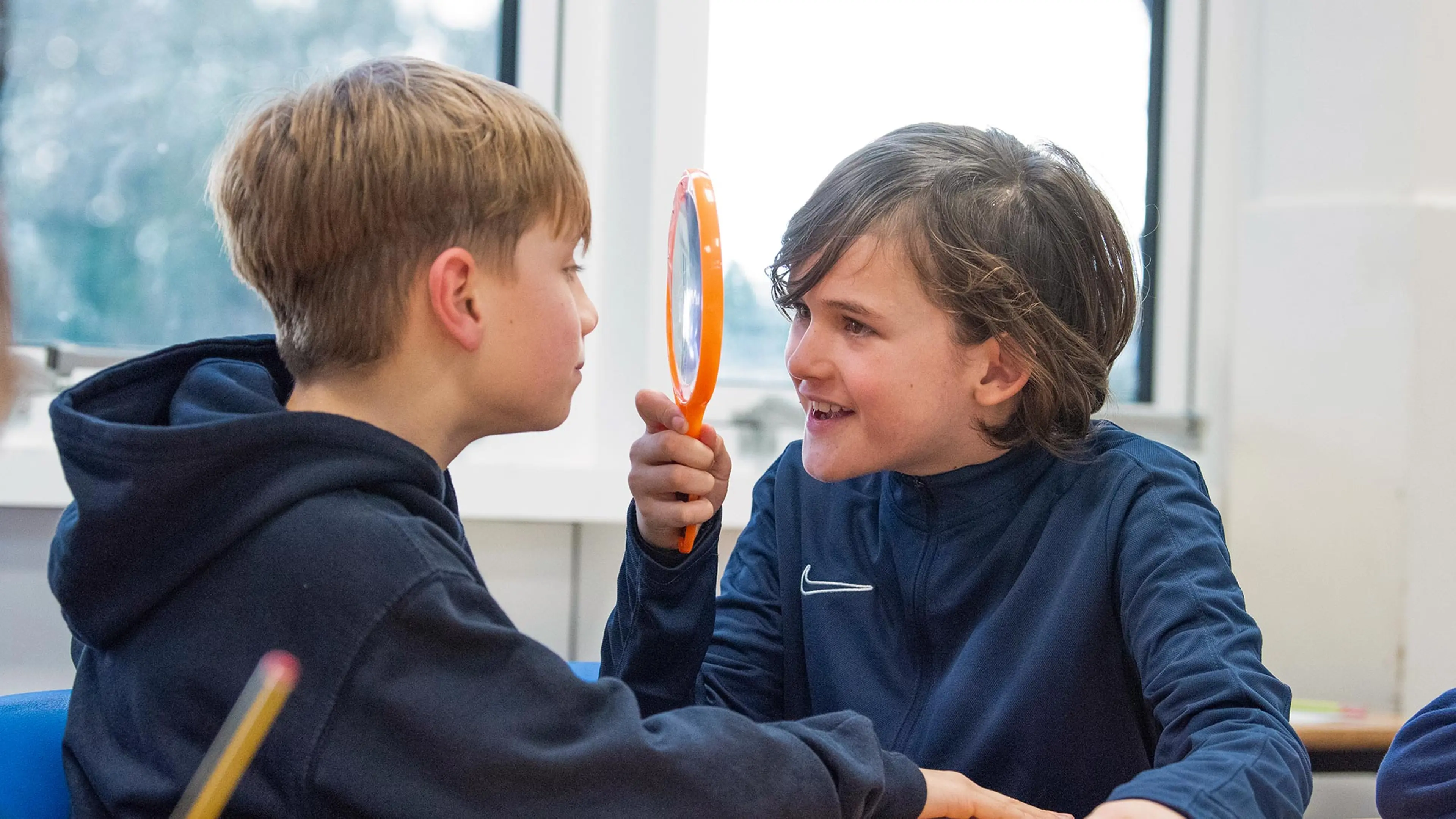 A schoolboy looking through a magnifying glass at his classmate.