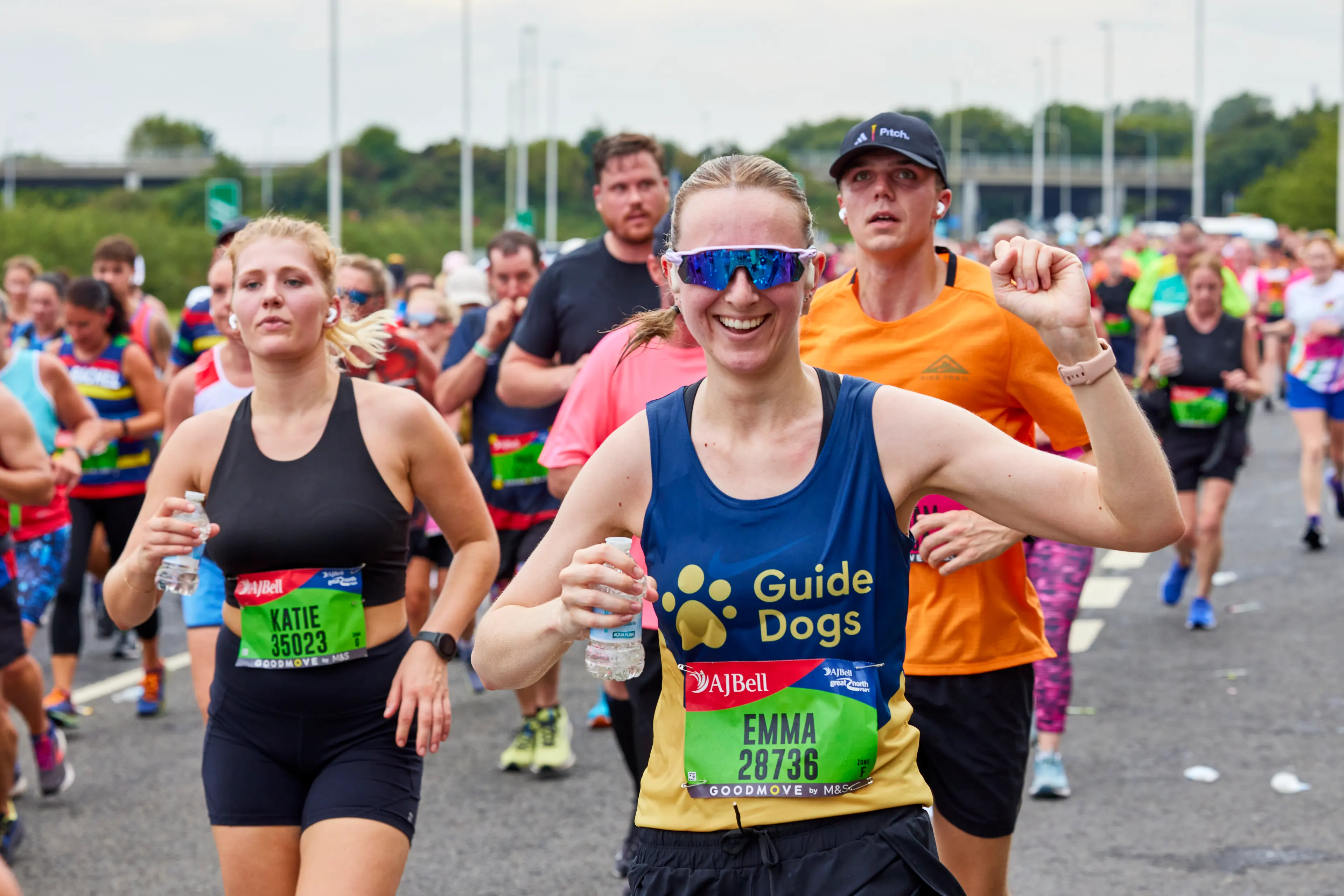 A runner in a race wearing a Guide Dogs vest smiles at the camera and waves her arm in the air.