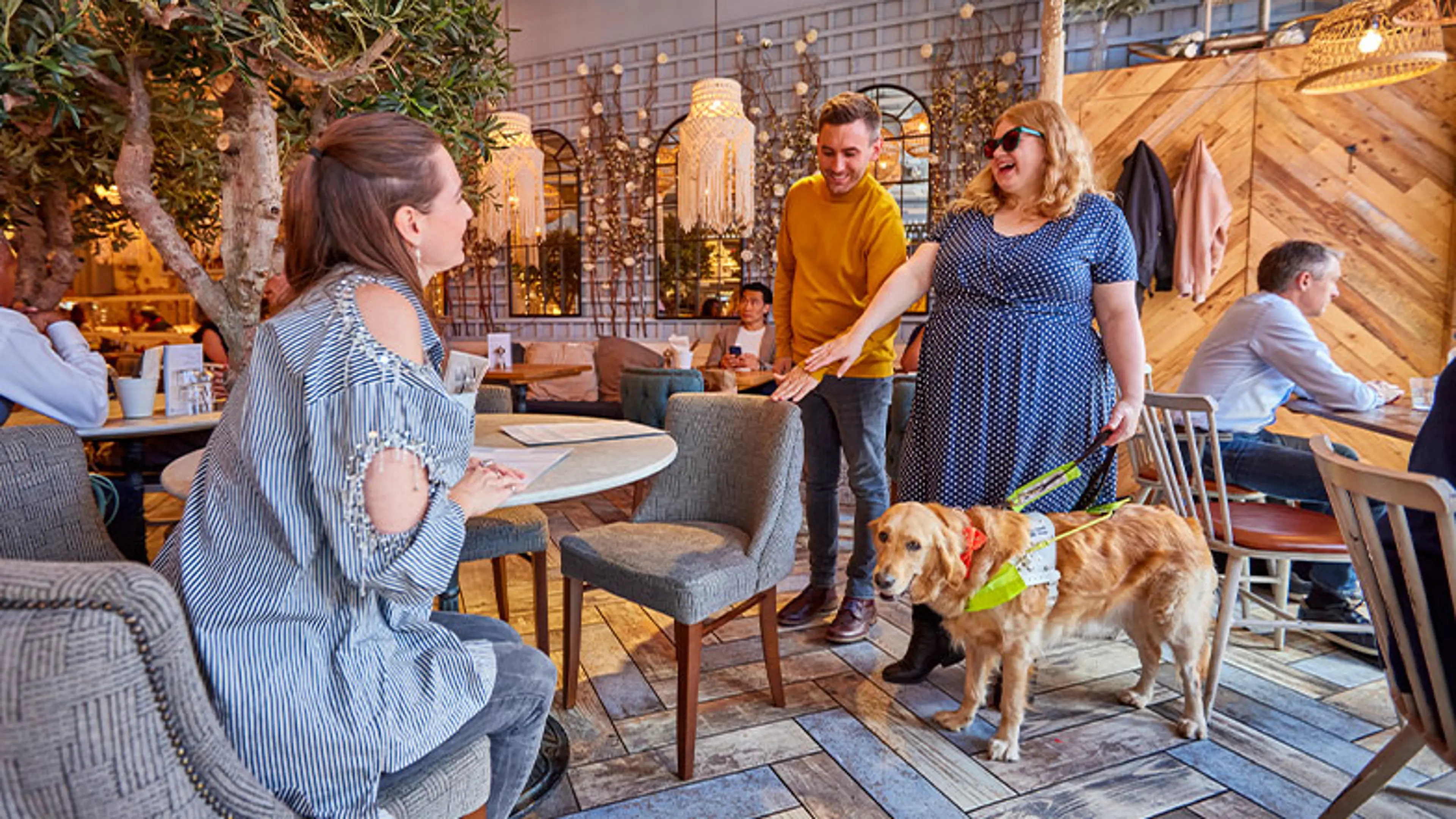 A man guiding a woman to find her chair in a café, by placing his hand on the back of the chair