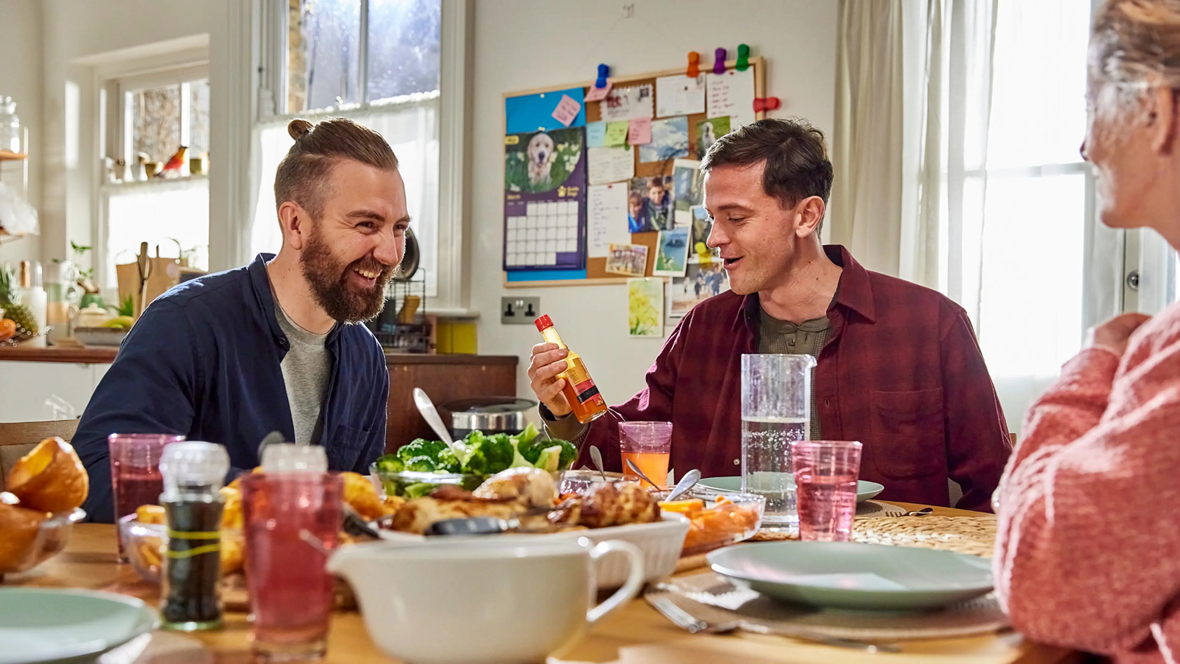 Two brothers sit at a table laughing together, one of the brothers holds a bottle of hot sauce he's used in a prank.