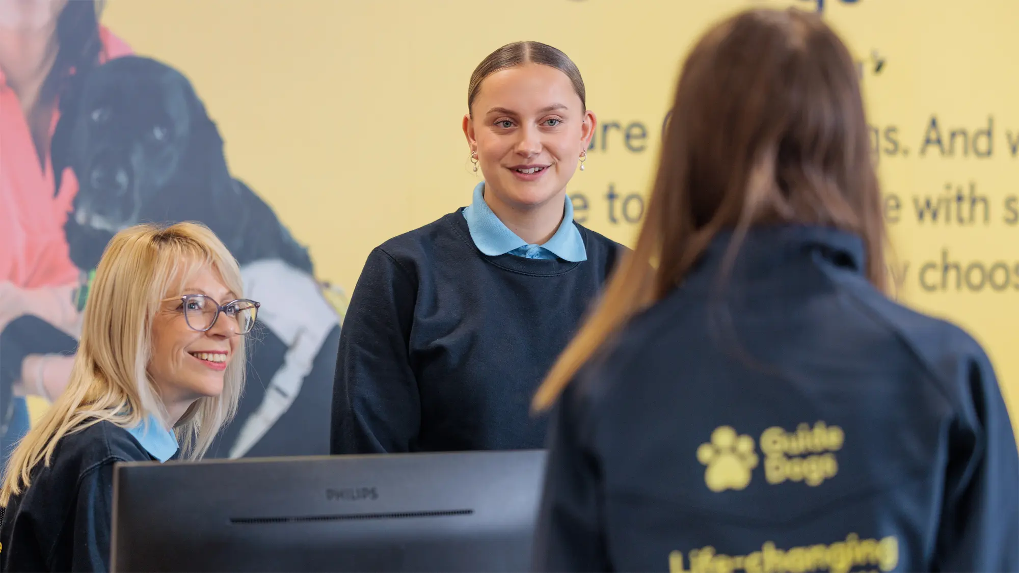 Two Guide Dogs staff members sit behind the reception desk. They talk to another colleague who has arrived at reception.