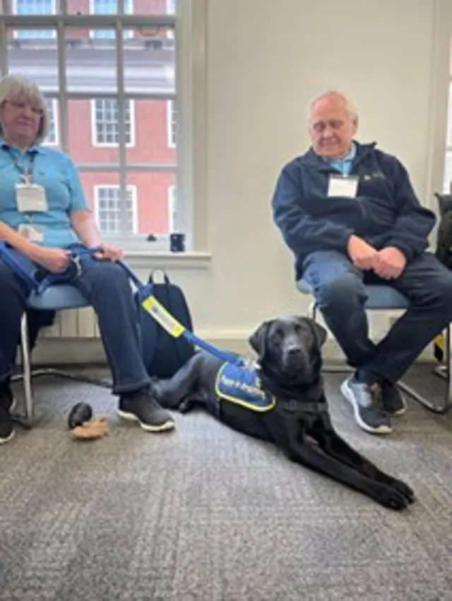 Black labrador, Dusty, is lying down and looking towards the camera while wearing a ‘puppy in training’ jacket, with her puppy raisers Sue and John sat on chairs next to her