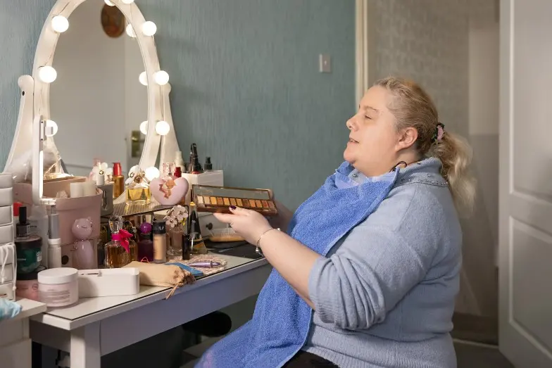 Marie, a woman with sight loss sits at her dressing table holding an eyeshadow palette. Her table has many beauty products on it and large lights around the mirror.