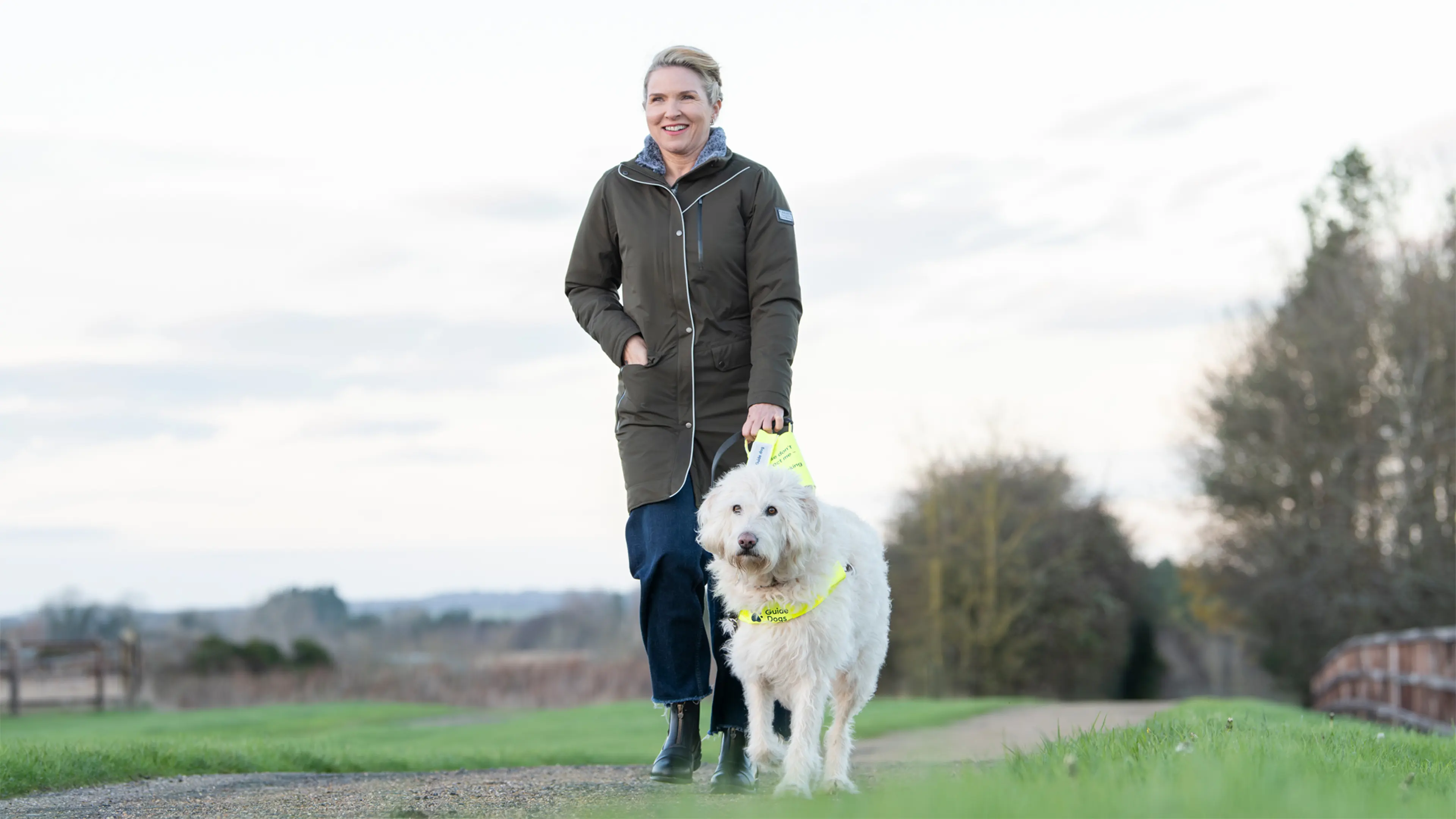 Guide dog owner Verity walks through a field holding the harness of her Labradoodle guide dog Luna.