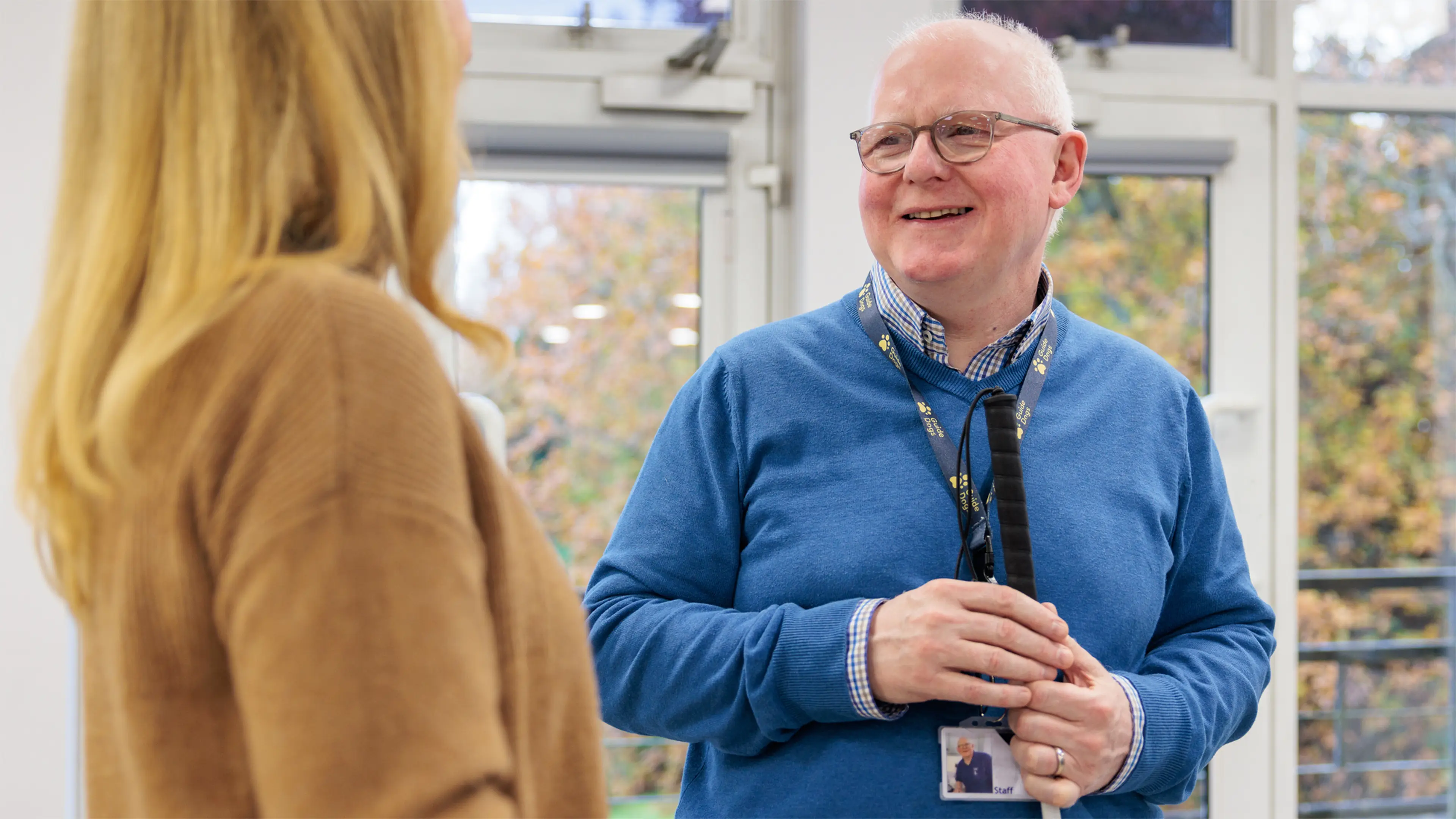 A Guide Dogs staff member smiles to another colleague as they stand by the office windows. He holds his cane as they speak.