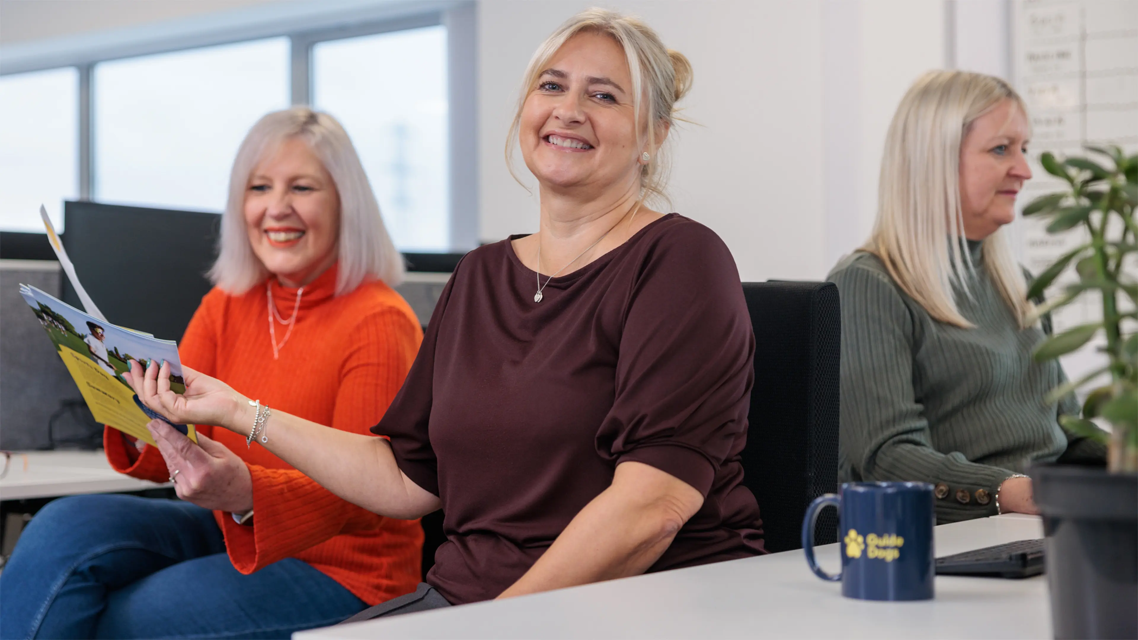A Guide Dogs staff member hands some fundraising posters to a colleague at the desk behind her. 