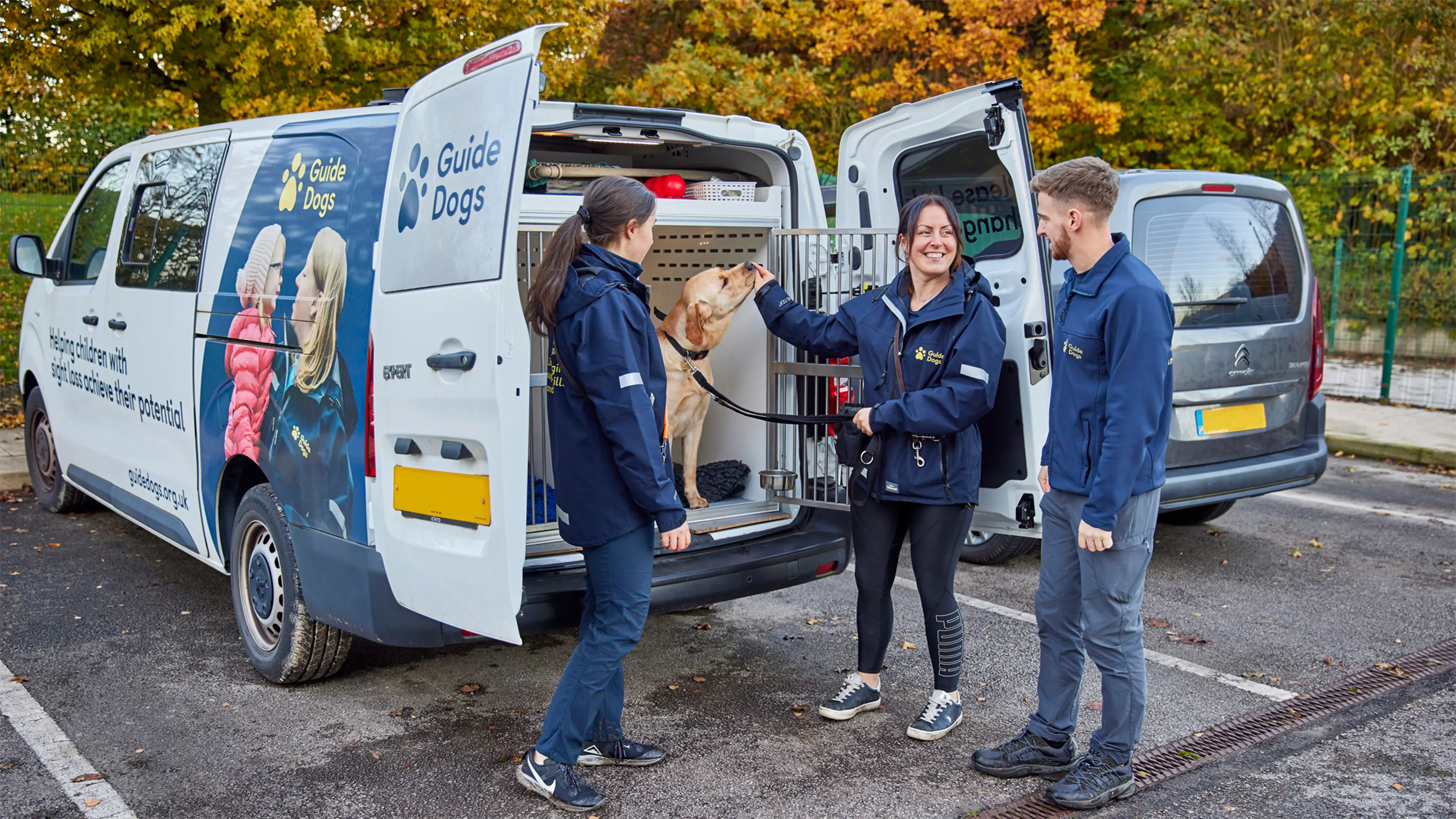 Three Guide Dogs Academy staff members stand by a parked Guide Dogs branded van. One of the staff members feeds a guide dog in training a treat.