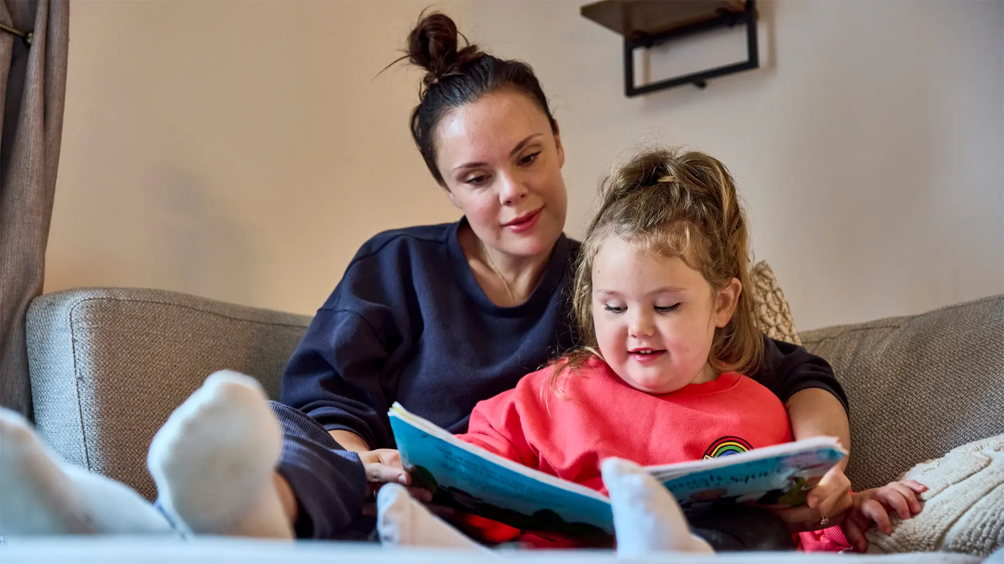 Mabel, a young girl with vision impairment, and her mum read a story together on the sofa. 